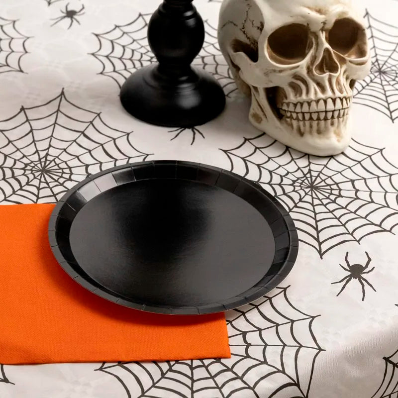 A black party plate and orange napkin sitting on a white tablecloth with a black spiderweb pattern, next to a decorative skull.