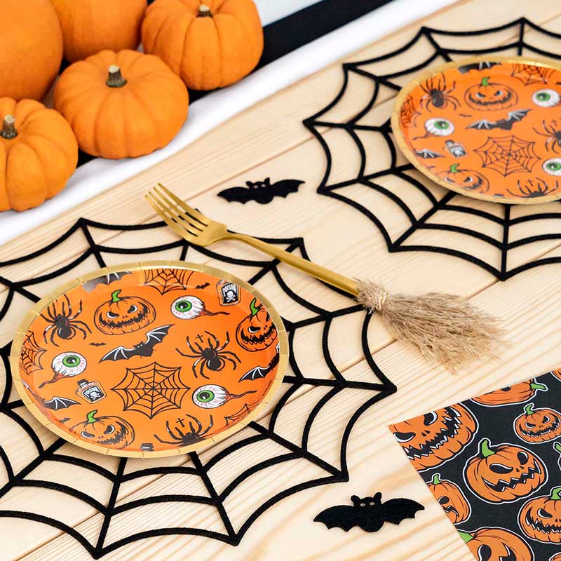 Halloween-themed table setting with orange plates and napkins printed with jack-o'-lanterns, spiders, bats, and eyeballs, arranged on a wooden surface with black spiderweb placemats, a gold fork, and small pumpkins in the background.