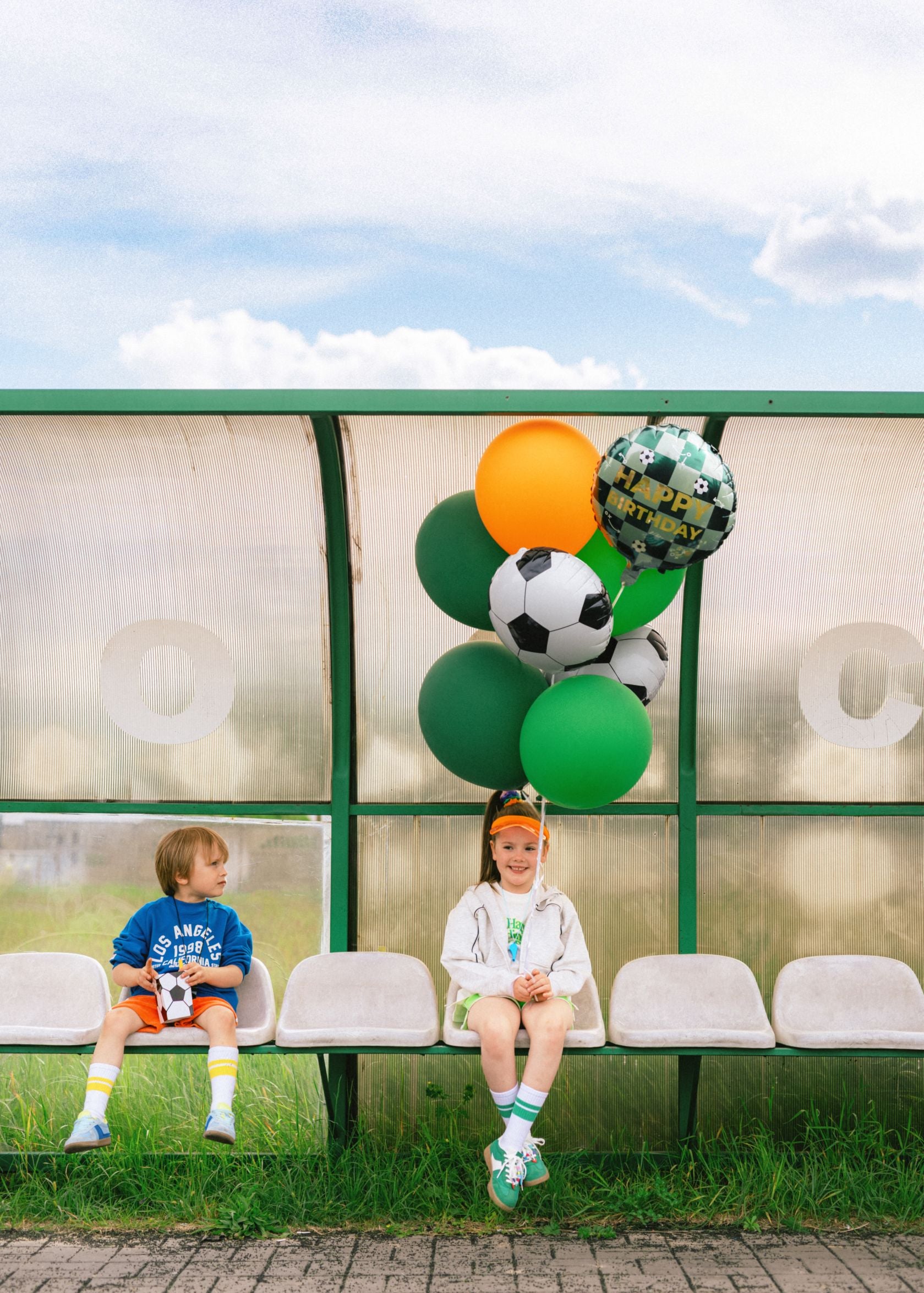 Two children sitting on white stadium bench seats outdoors, with a bunch of balloons next to them. The balloon bunch includes a checkered "Happy Birthday" foil balloon with soccer balls, solid green balloons, a solid orange balloon, and black and white soccer ball-patterned balloons. The background shows a light blue sky with clouds and a green grassy area.