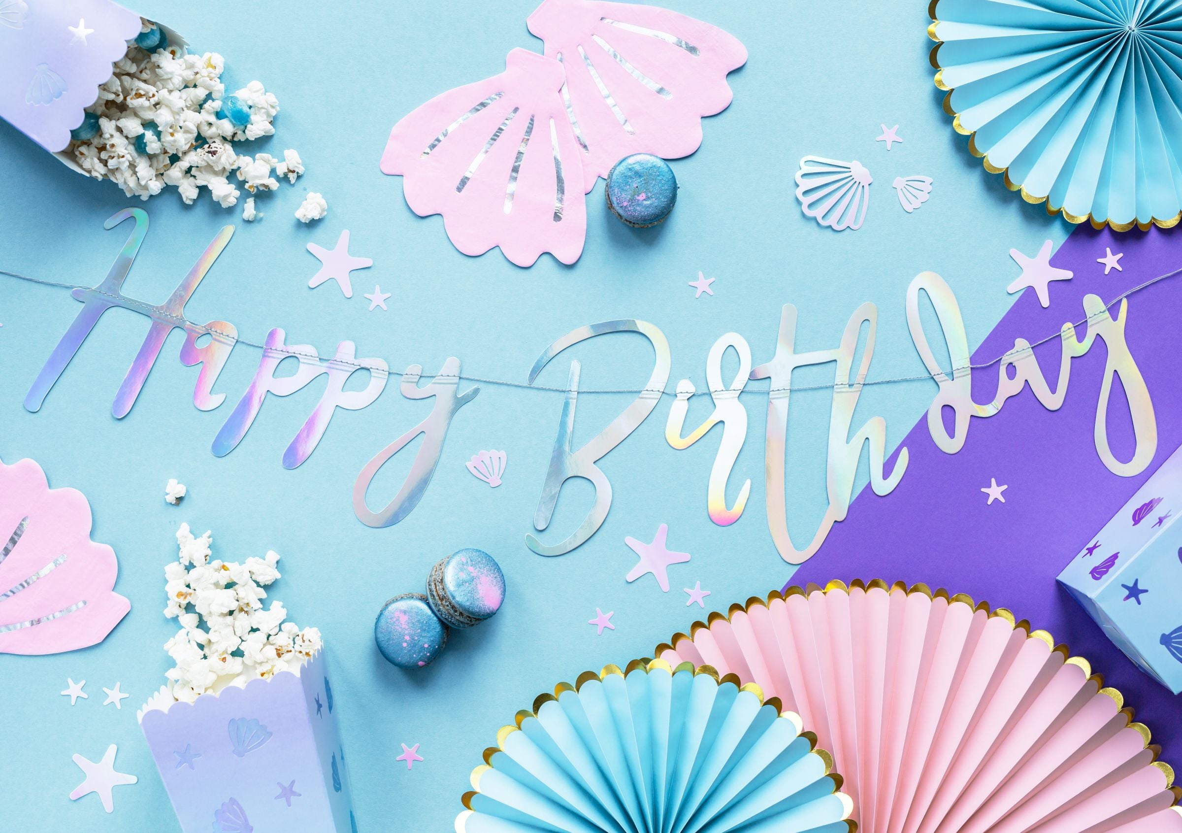 An overhead flat lay of iridescent mermaid-themed party supplies on a light blue and purple background, featuring the "Happy Birthday" iridescent banner, pink seashell cutouts, popcorn boxes, iridescent confetti, and blue and pink paper fan decorations.