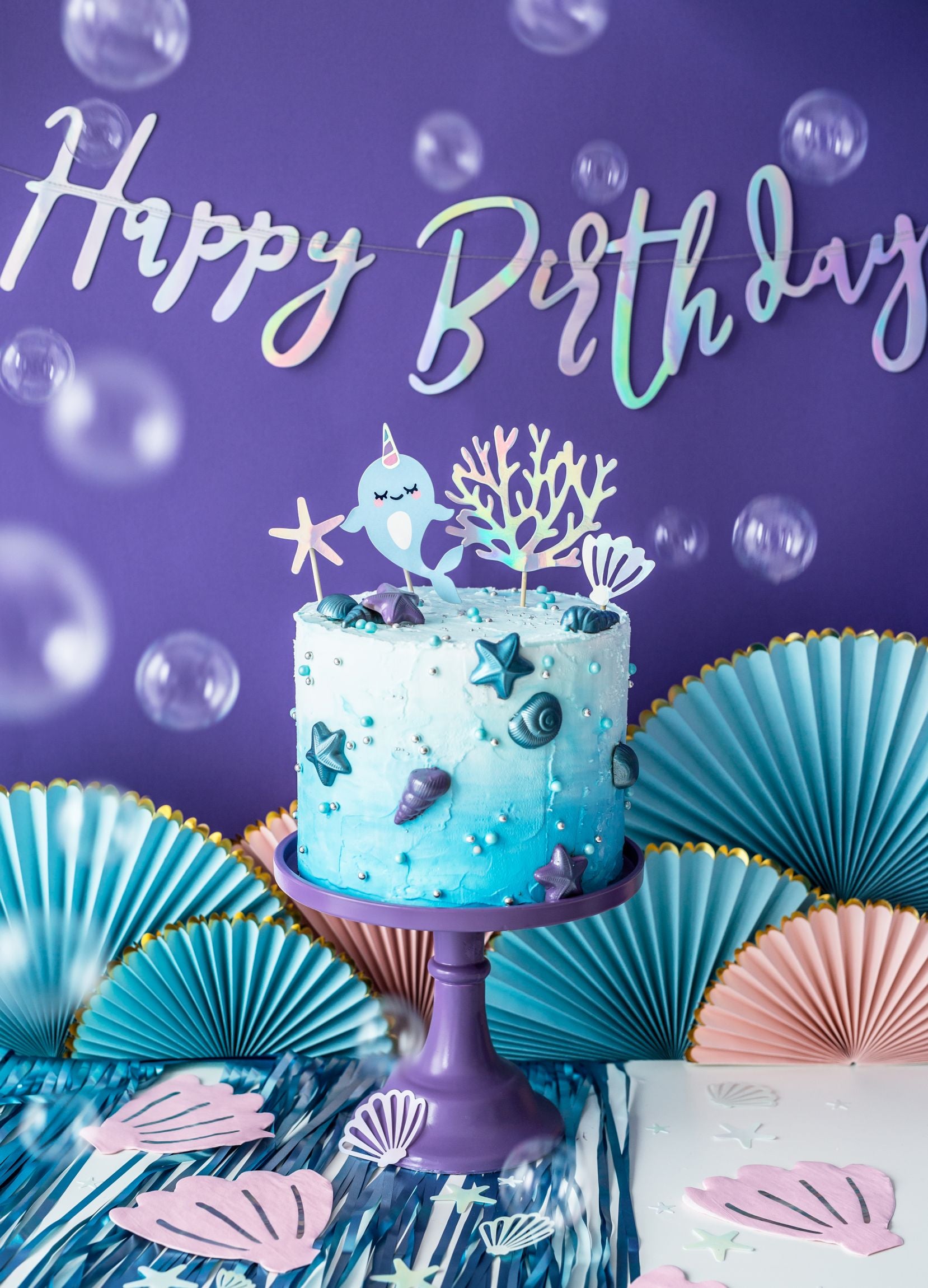 A vibrant birthday party setup with a shimmering iridescent "Happy Birthday" banner hanging against a deep purple wall. Below it, a two-tiered ombre blue and white cake adorned with sea creatures and stars sits on a purple cake stand, surrounded by blue and pink paper fan decorations and clear bubbles.
