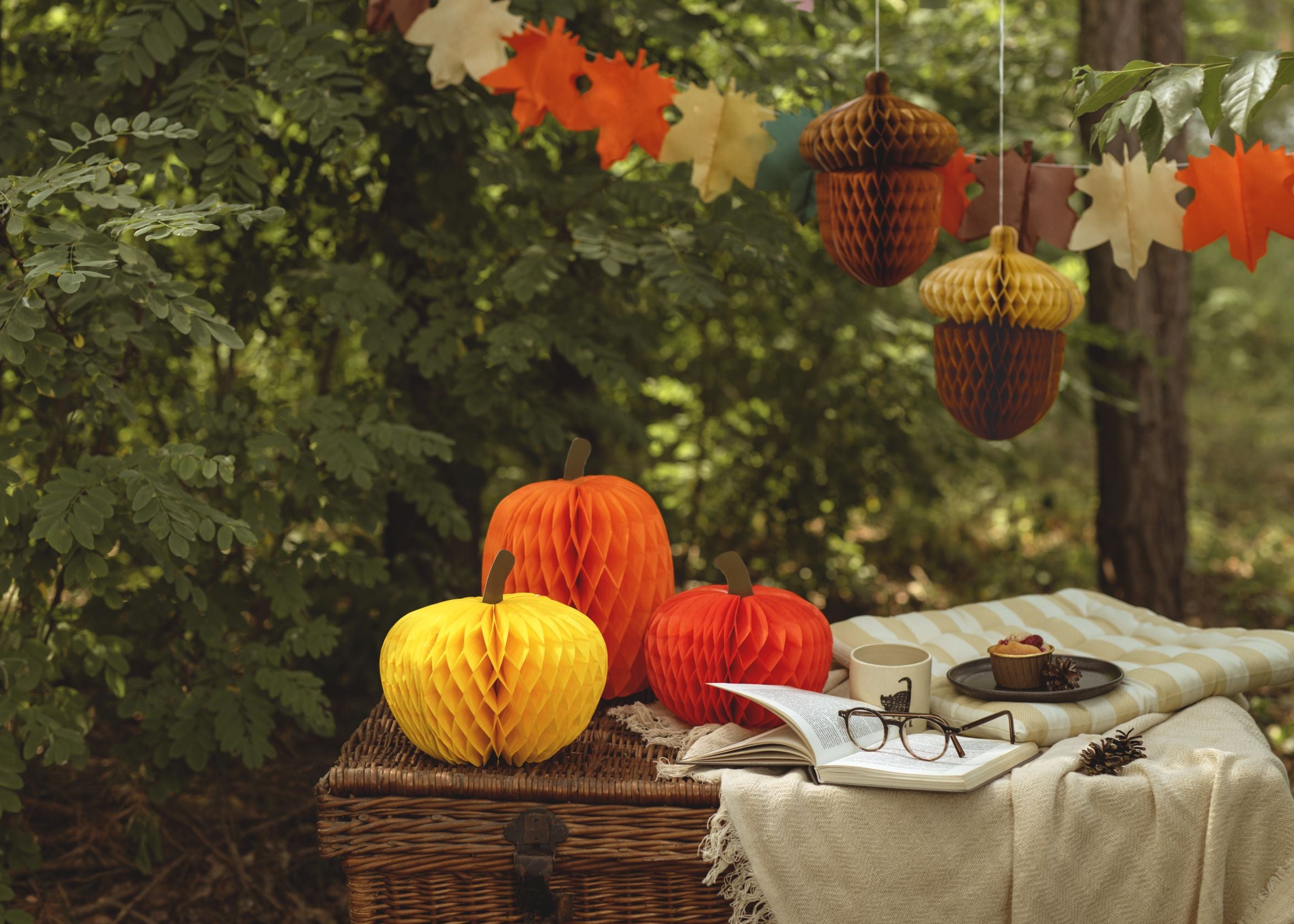  An outdoor autumn scene with three honeycomb paper pumpkins (orange, yellow, red) displayed on a woven basket. Next to them, a blanket-covered table holds a book with glasses, a mug, and a small cake. In the background, a garland of autumn leaves and two hanging honeycomb acorn decorations are visible amongst green trees.