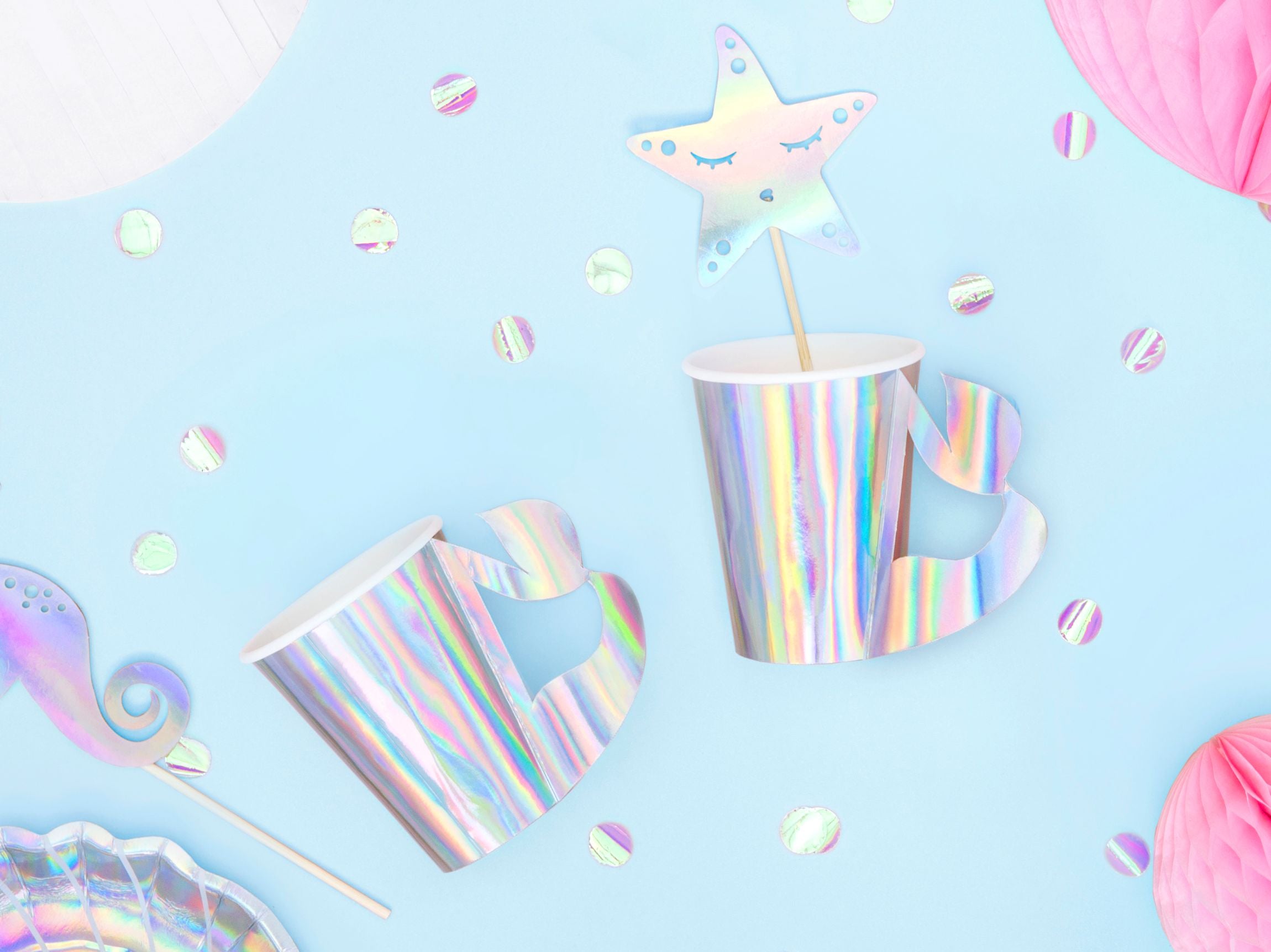 An overhead flat lay of iridescent mermaid-themed party supplies on a light blue background, featuring two shimmering cups with mermaid tail handles, one containing a star-shaped topper, alongside iridescent confetti, a matching plate, and pink honeycomb decorations.