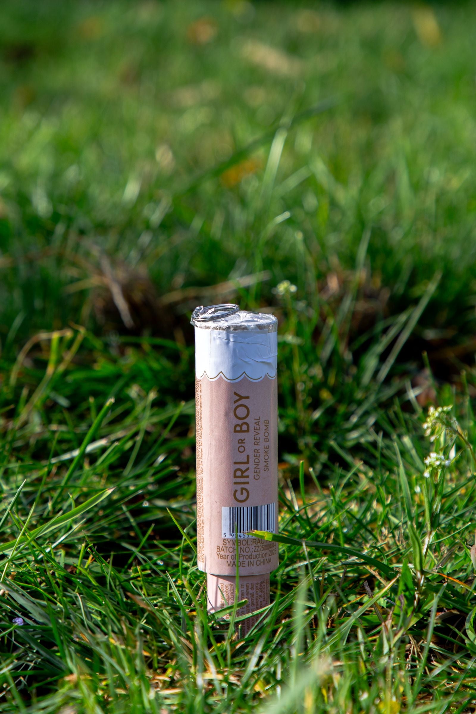 A gender reveal smoke bomb standing unlit in green grass, featuring pastel packaging with "GIRL OR BOY" text.