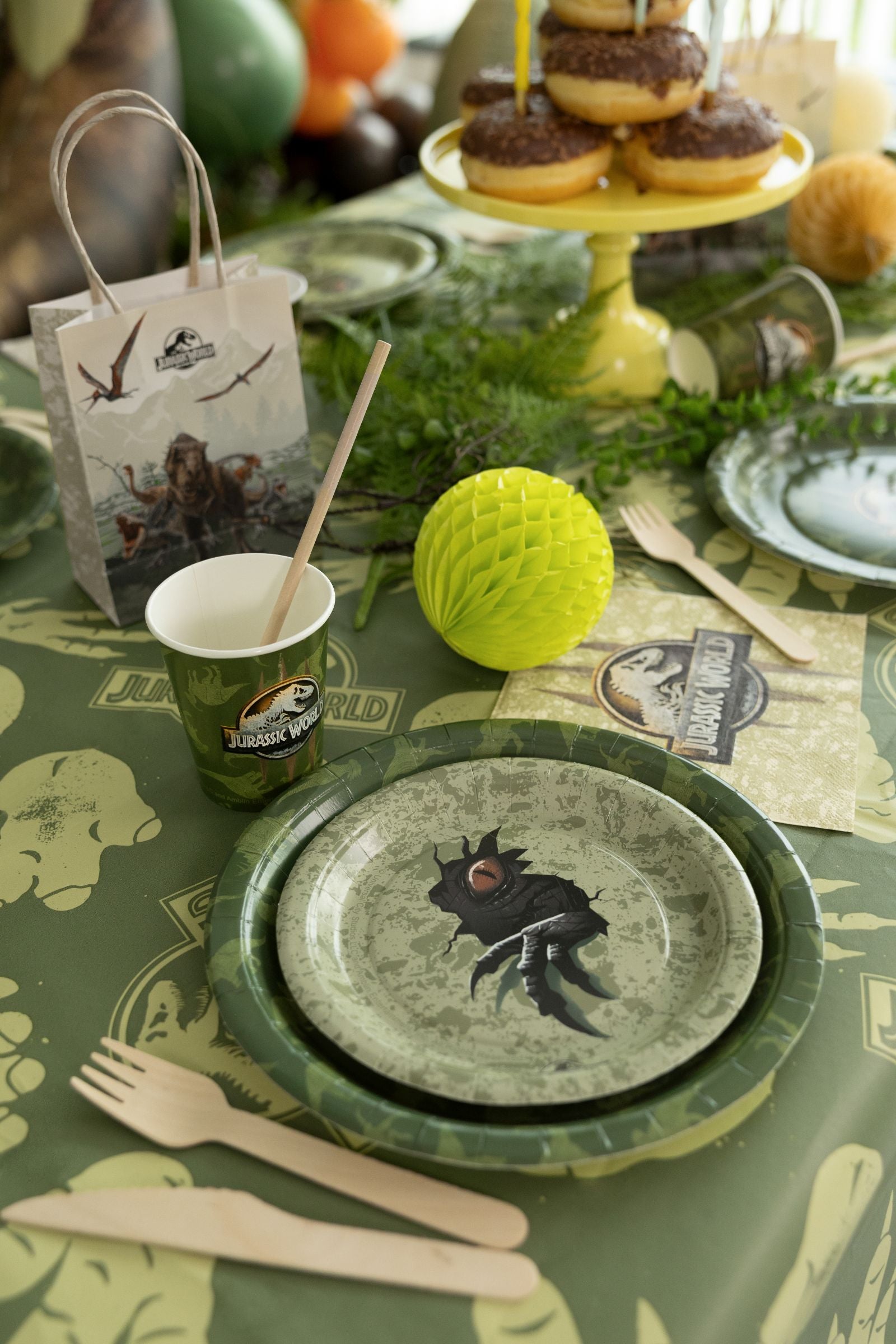 Jurassic World themed party table with camouflage dinosaur plates, character cups, napkins, and wooden cutlery.