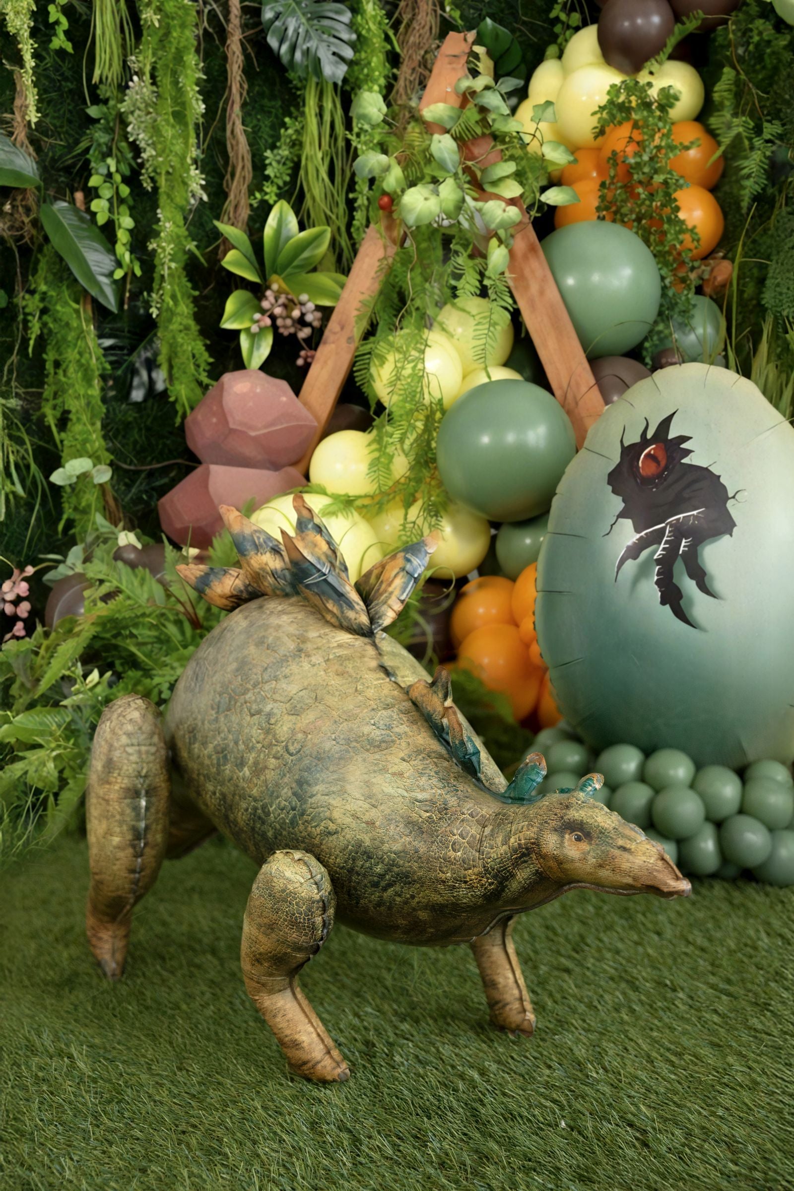 Giant 3D standing Stegosaurus foil balloon on a grass floor in front of a jungle-themed birthday backdrop with greenery and dinosaur eggs.