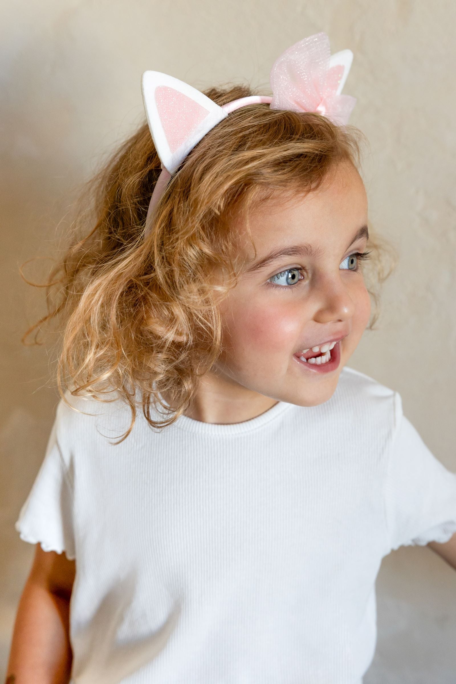 A close-up of a young child with a bright, happy expression, wearing a pink headband with white cat ears and a pink organza bow, against a textured beige background.