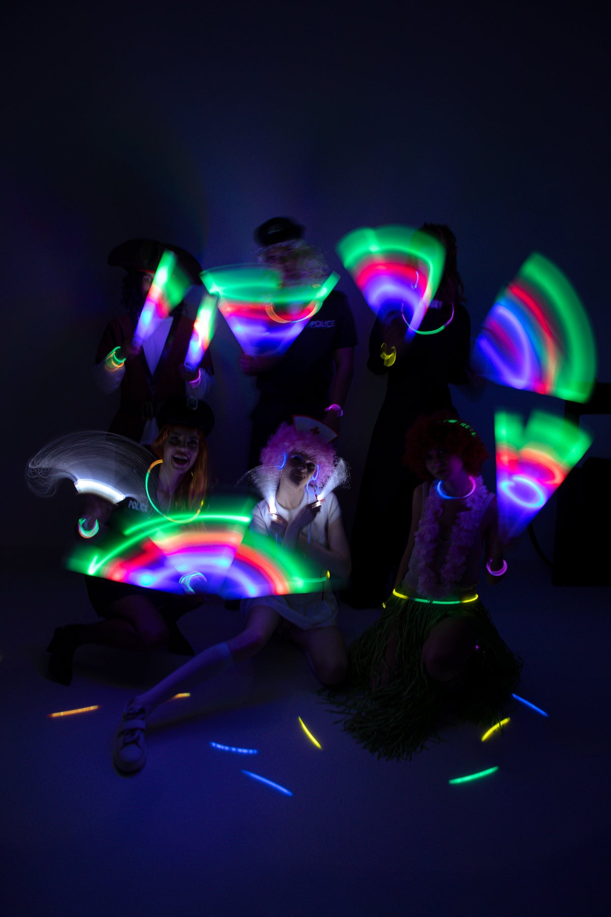 A group of people posing in a dark room wearing and holding glowing neon UV-reactive accessories, including rainbow wristbands, necklaces, and handheld glow fans that emit green, pink, and blue light.