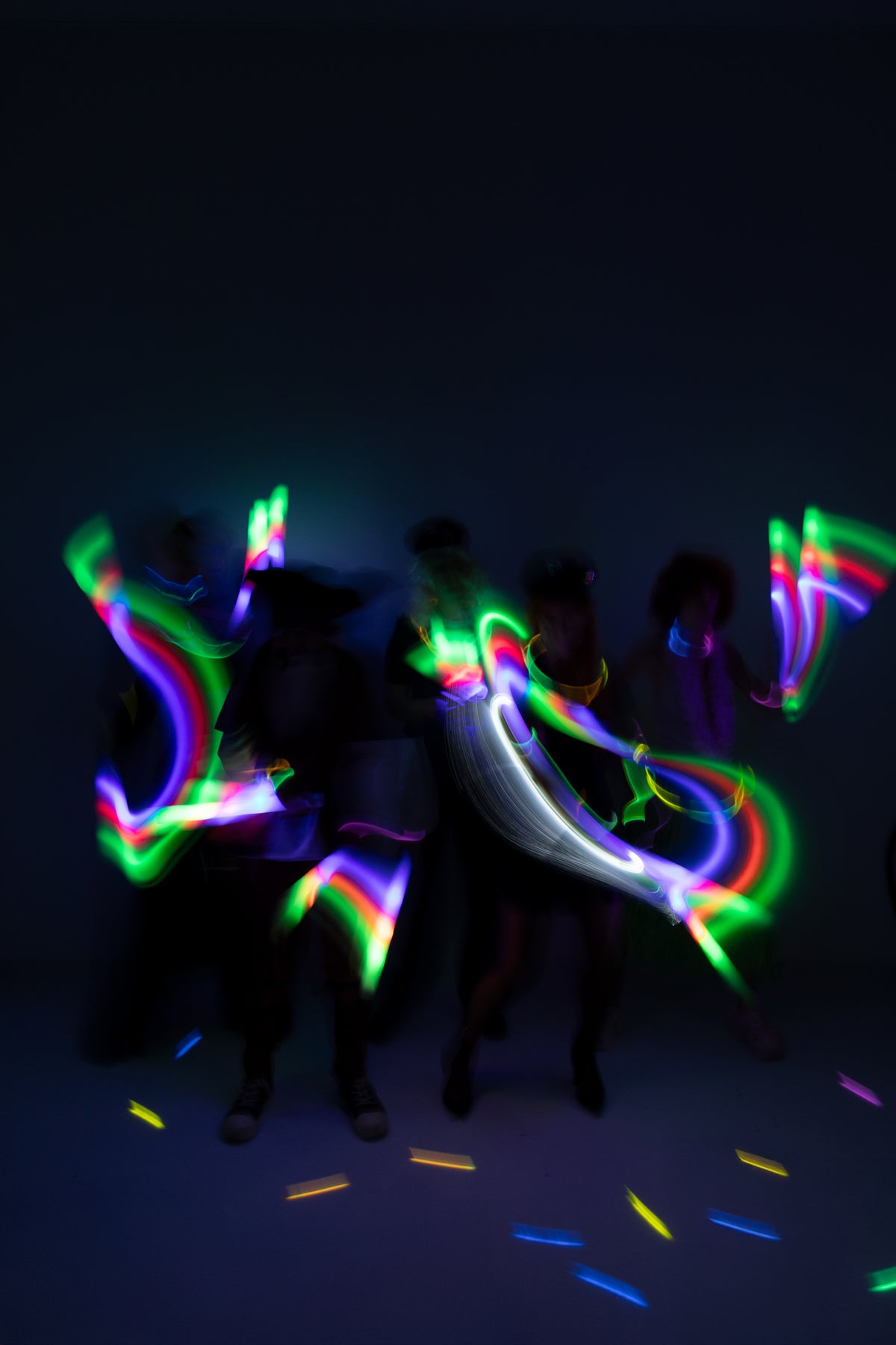 Several people dancing in a dark room while holding glow-in-the-dark rainbow light sticks, creating vibrant neon light trails in green, blue, purple, pink, and white.