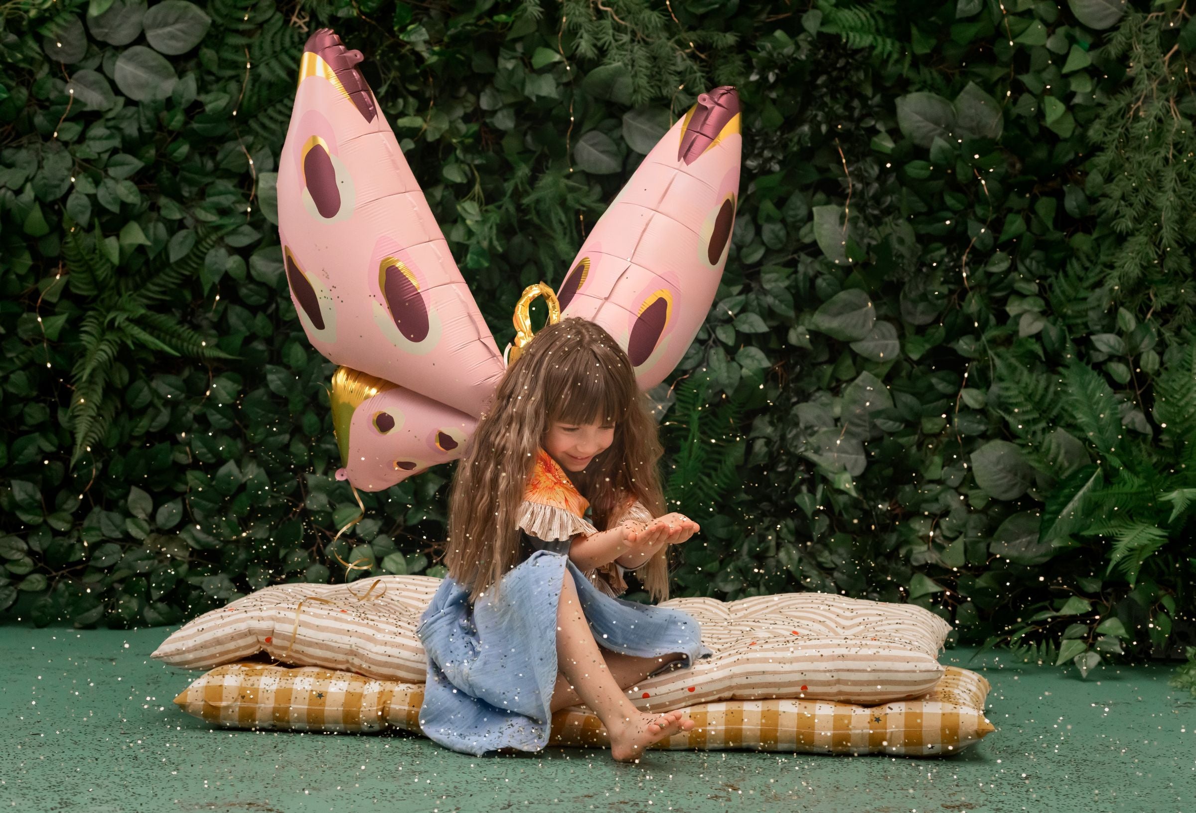 A child with long, wavy hair is seated on a stack of pillows, playing with confetti and looking down at their hands. A large pink and gold foil butterfly balloon is positioned behind them like wings, with a forest-themed backdrop.