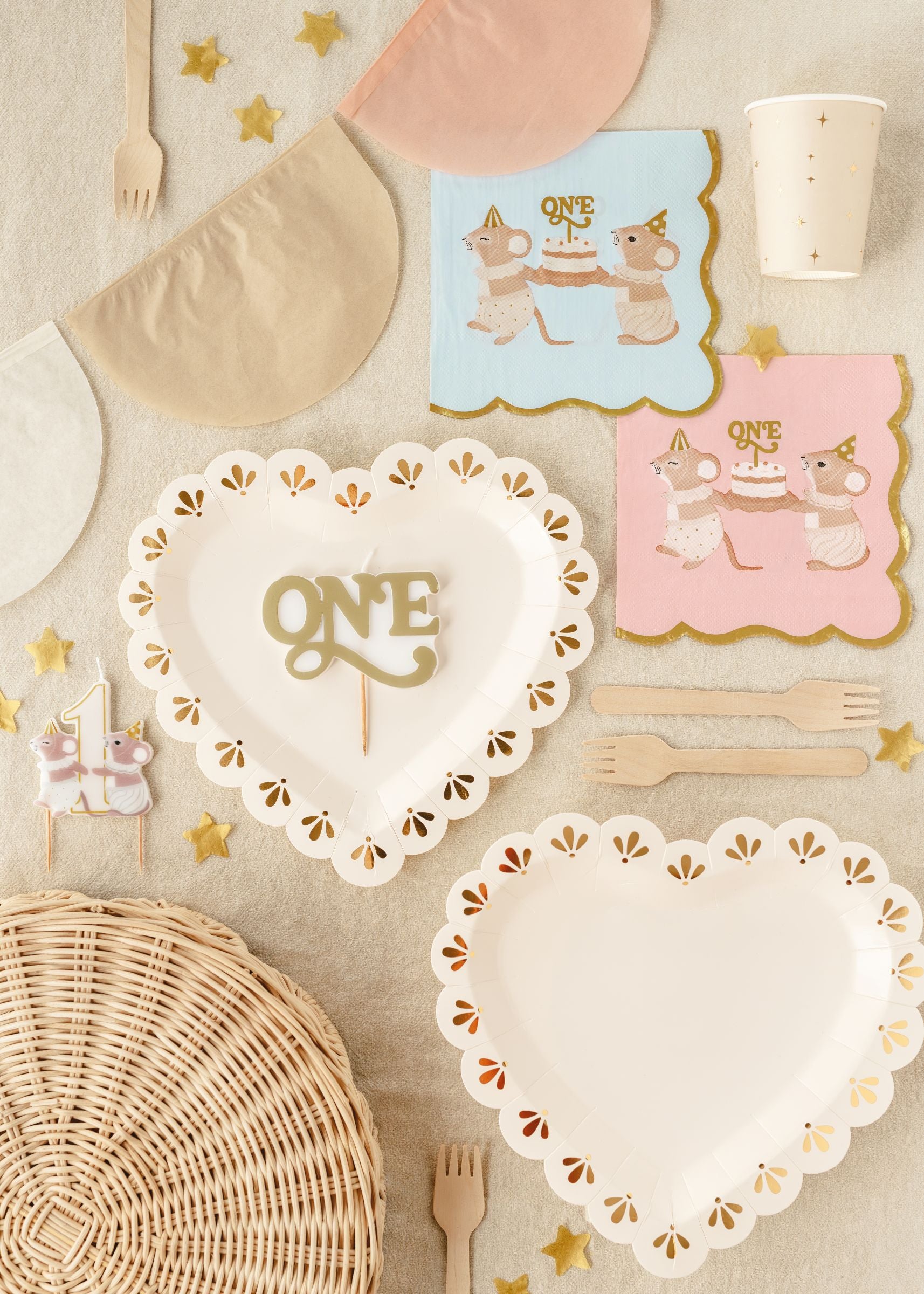 A flat lay party setup featuring a mice-themed number one birthday candle alongside white heart-shaped plates with gold foil details, pastel mice napkins, wooden cutlery, and gold star confetti.