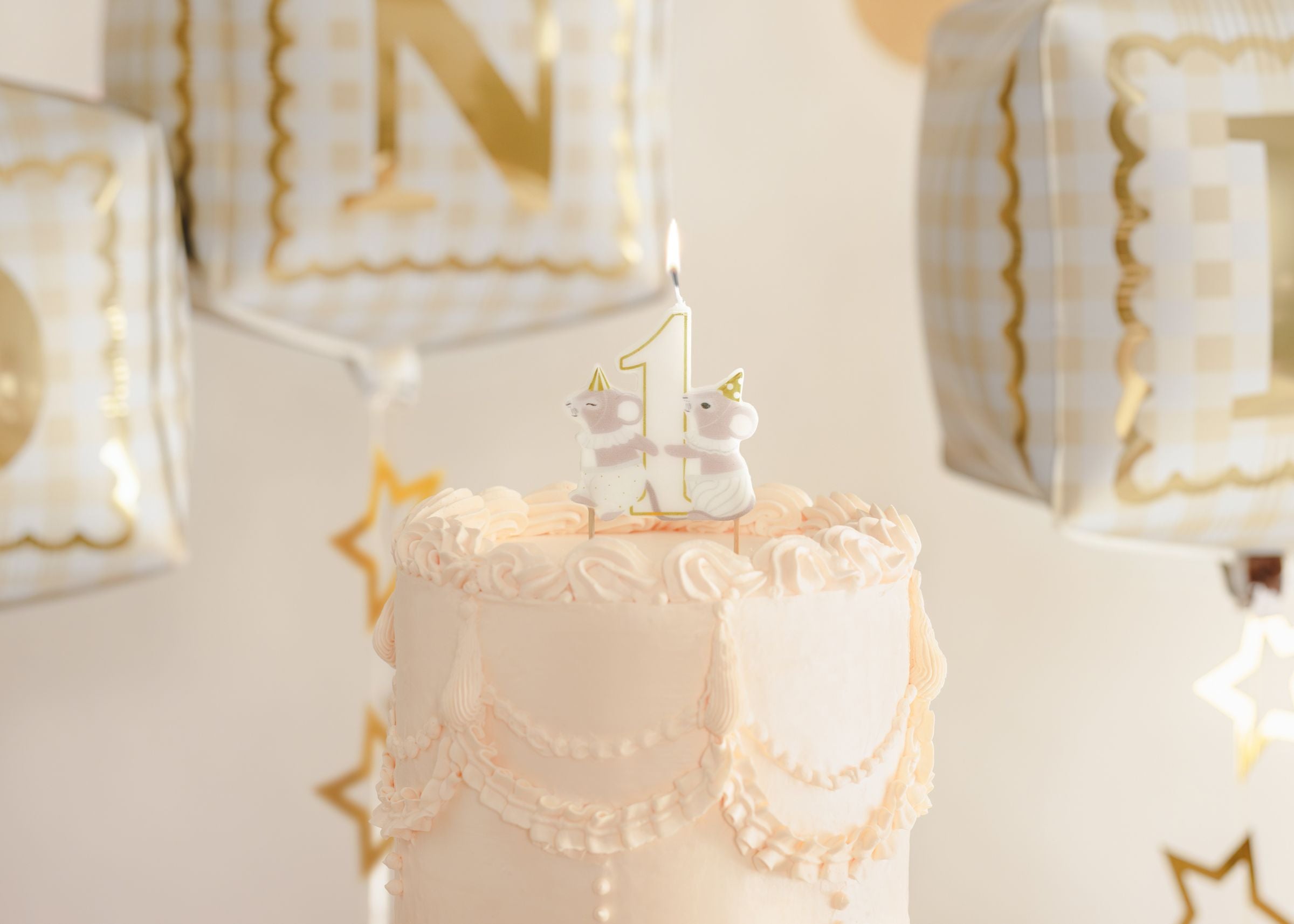 A beautifully piped pastel pink first birthday cake topped with a lit, mice-themed number one birthday candle, with foil balloons in the background.