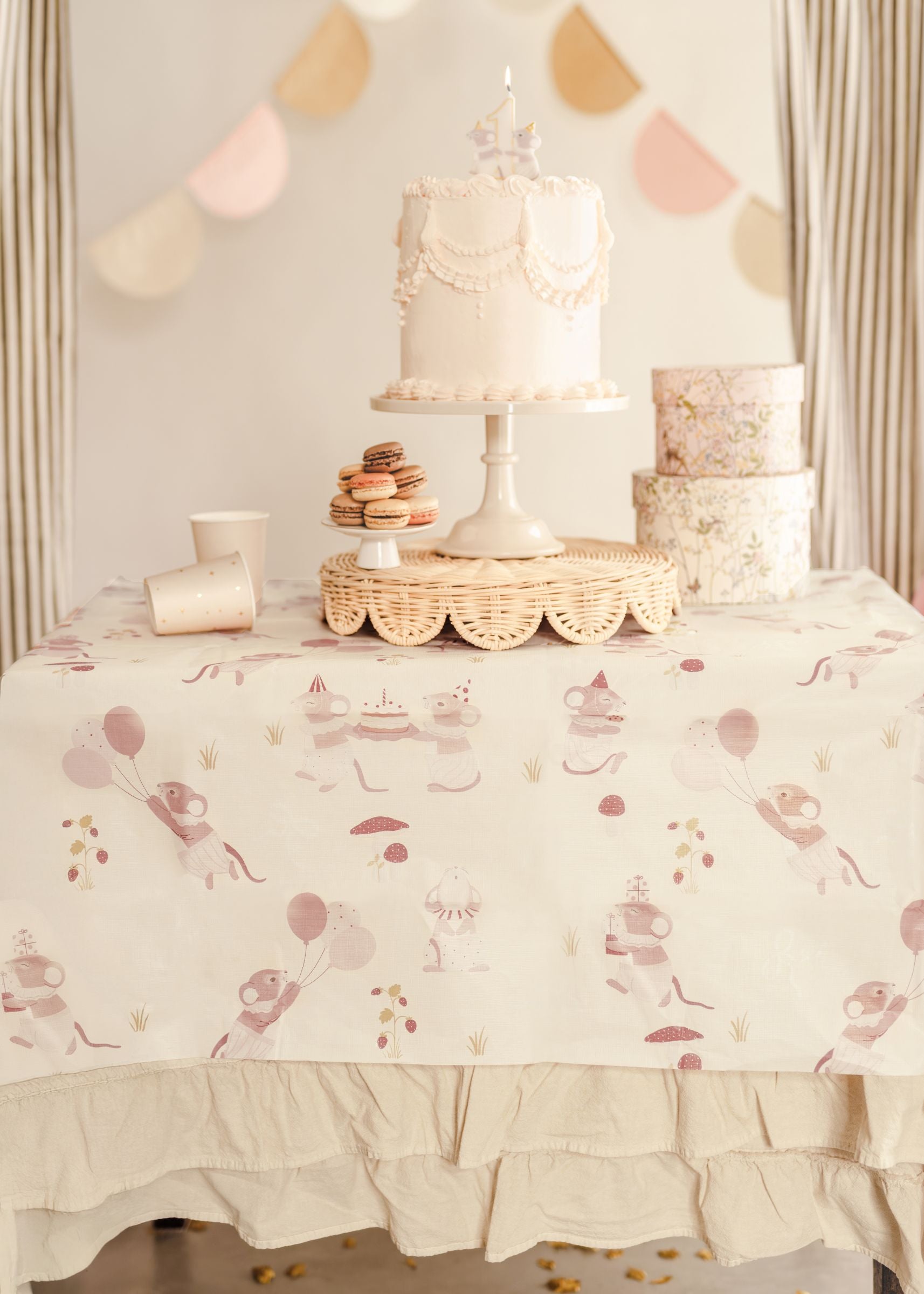 A styled party table covered in a beige mice-themed paper tablecloth, topped with a white birthday cake, macarons, and pastel cups.