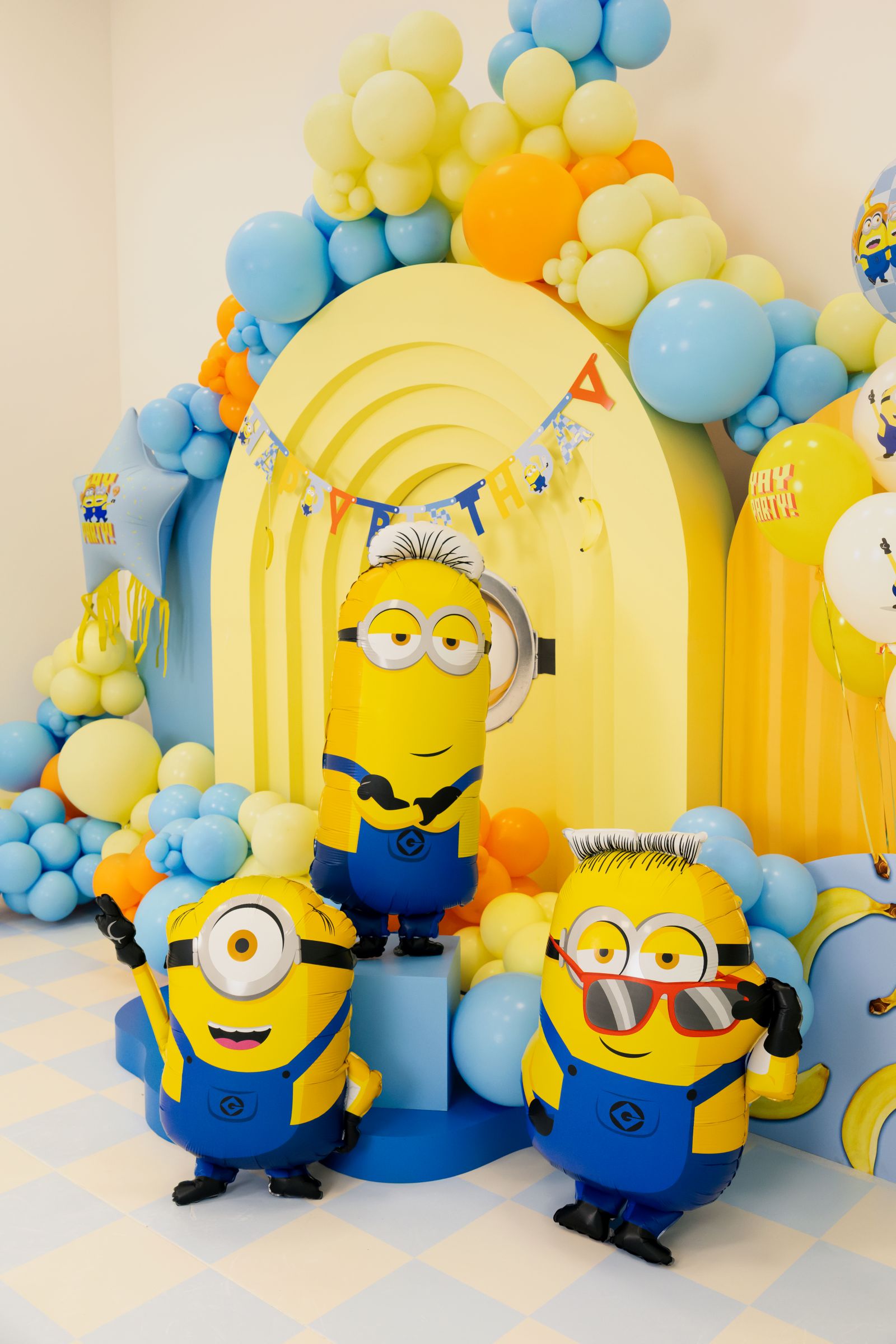 Giant Minion Ron foil balloon with red sunglasses displayed in a blue and yellow birthday party setting with Minion Mel and Minion Tim.
