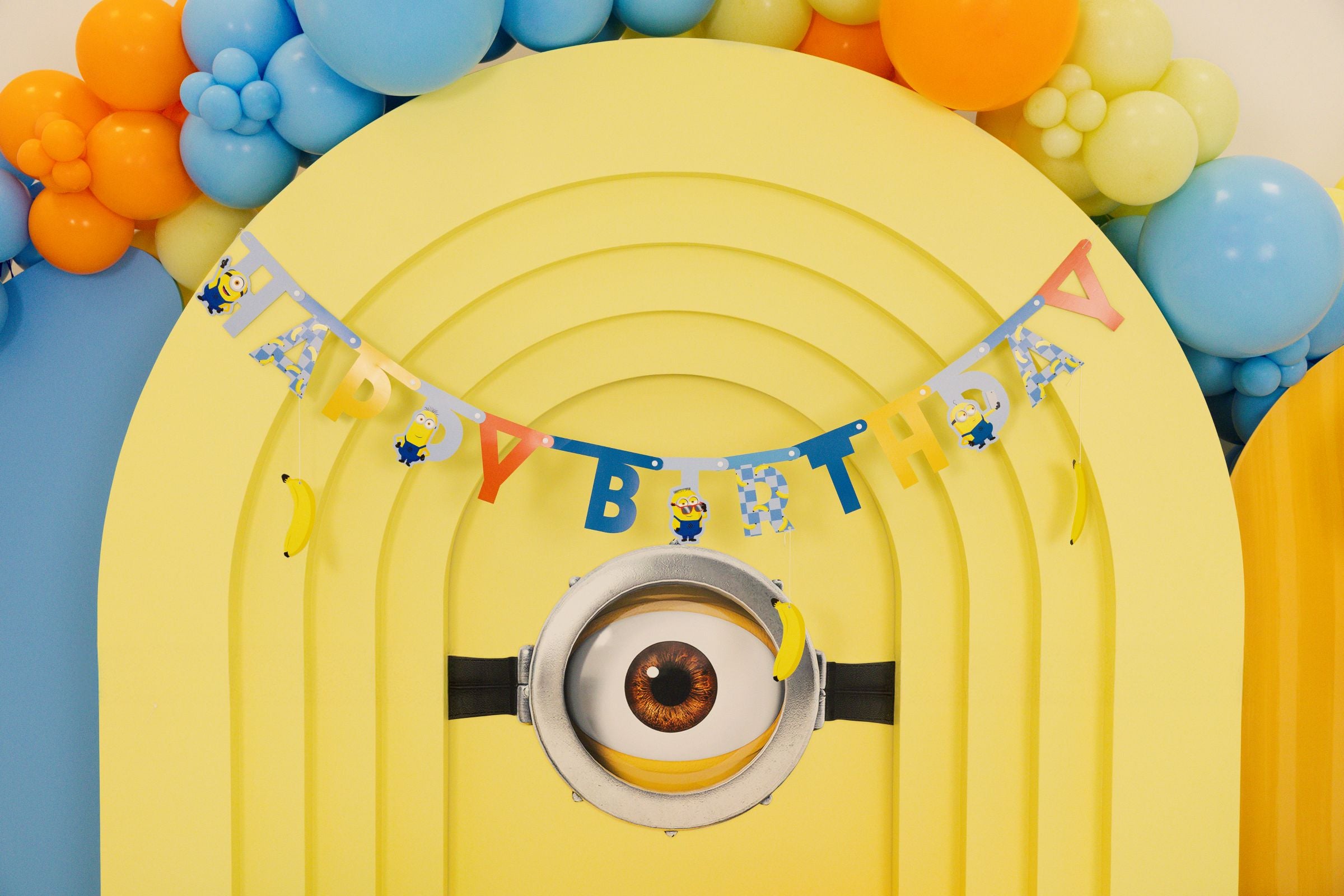 Minions Happy Birthday paper banner displayed across a yellow arched backdrop with a large Minion eye and colorful balloon arch.