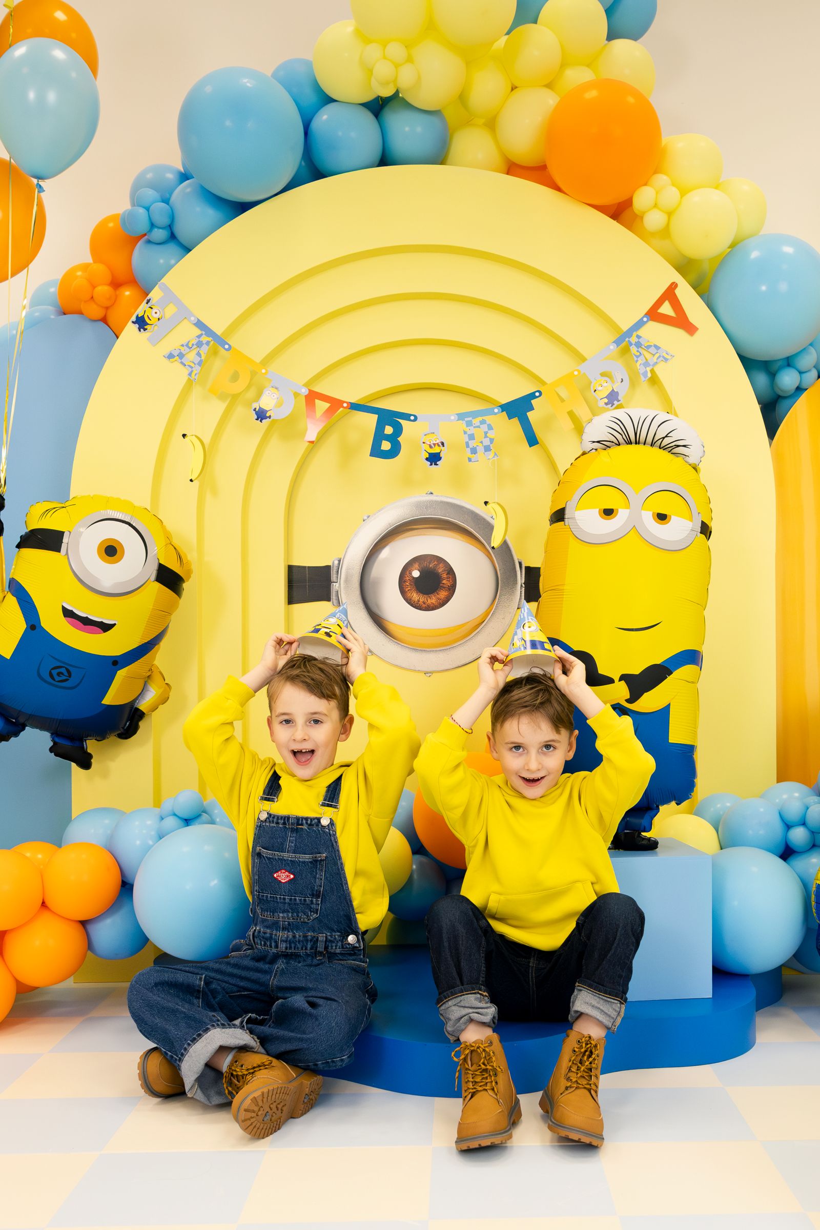 Two children posing in front of a birthday backdrop with Minion characters and balloons.
