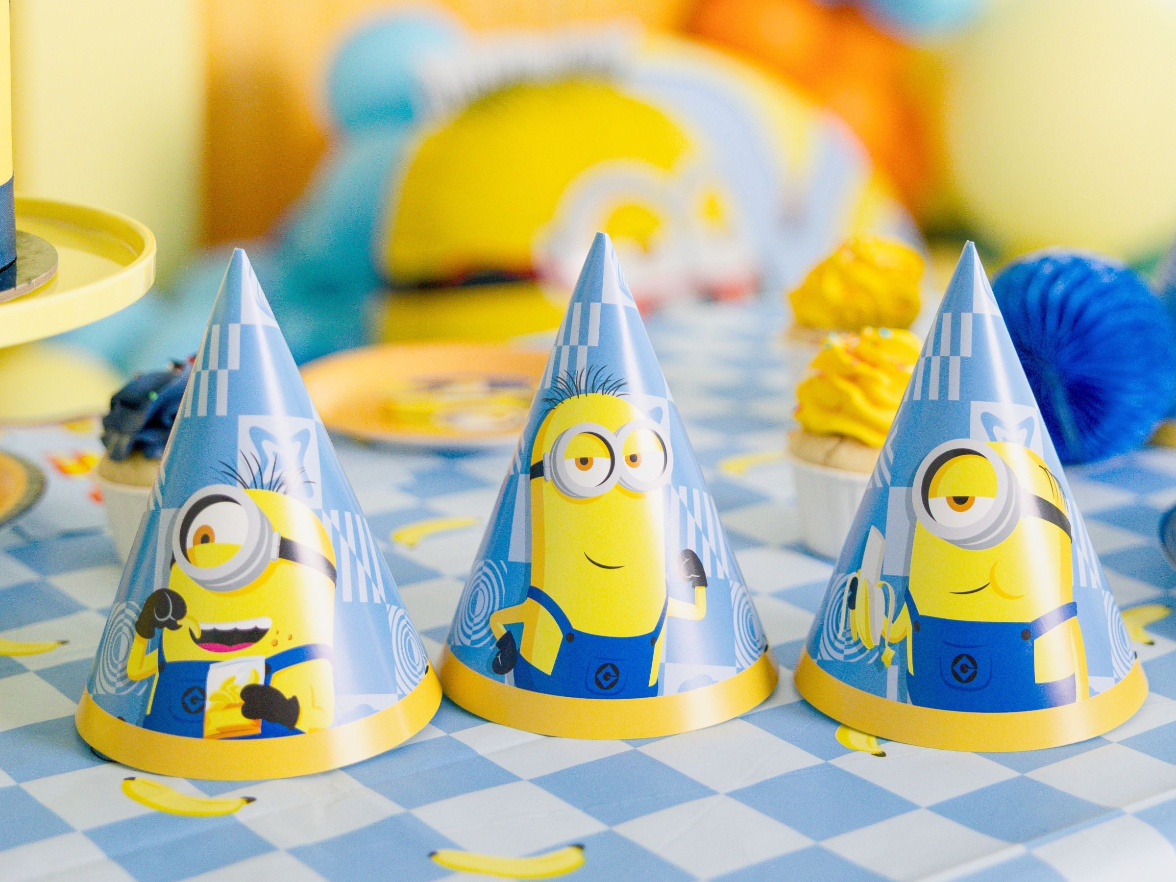 Three blue Minions themed party hats arranged on a matching checkered tablecloth next to yellow cupcakes.