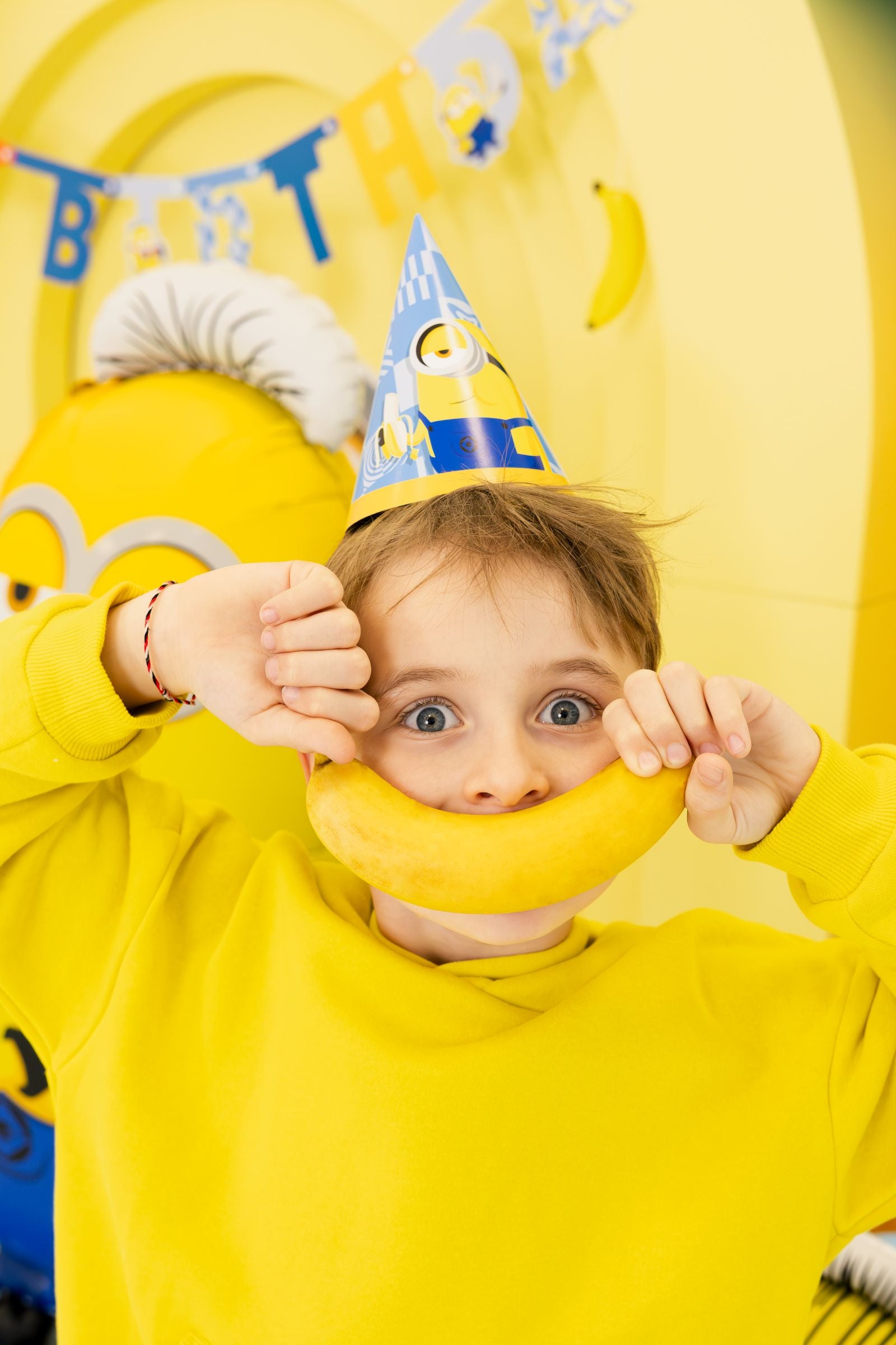 A playful party guest in a yellow jumper wearing a Minions cone hat, holding a banana like a big yellow smile.