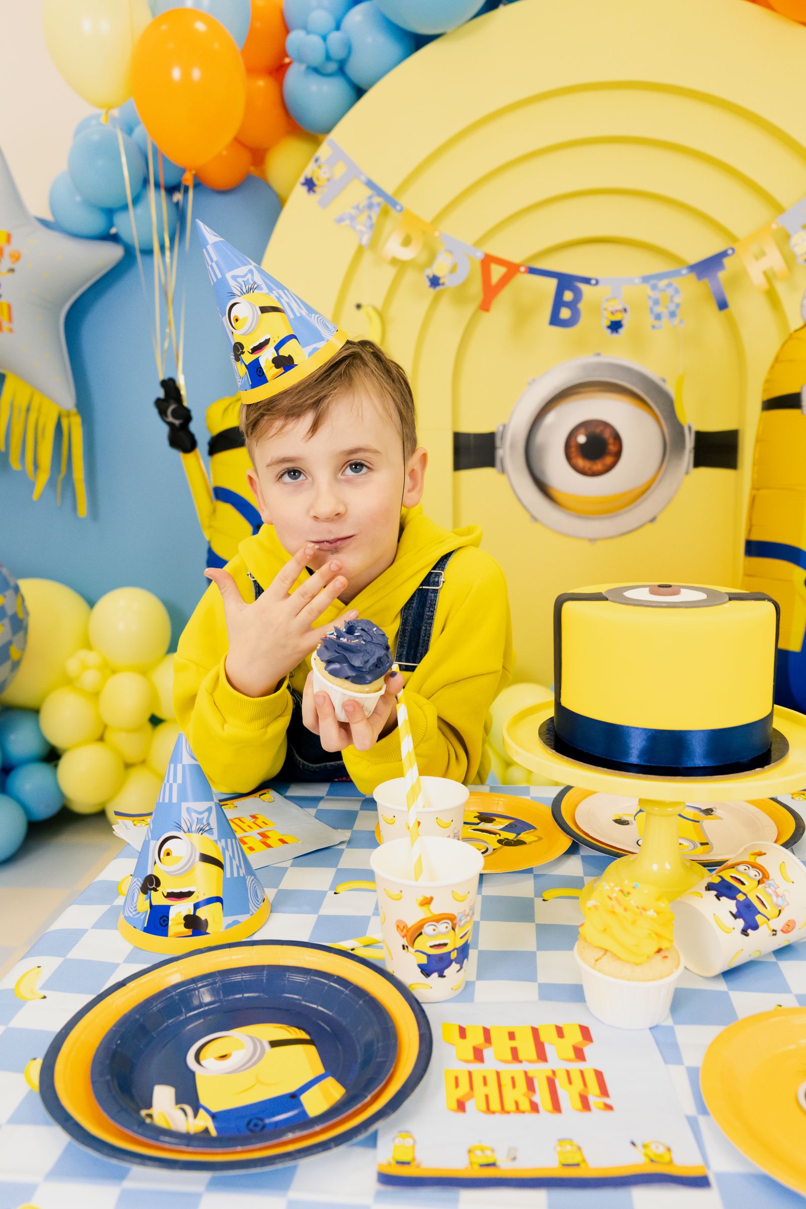 A young boy in overalls and a Minions party hat eating a blue cupcake at a fully decorated Despicable Me themed birthday table.