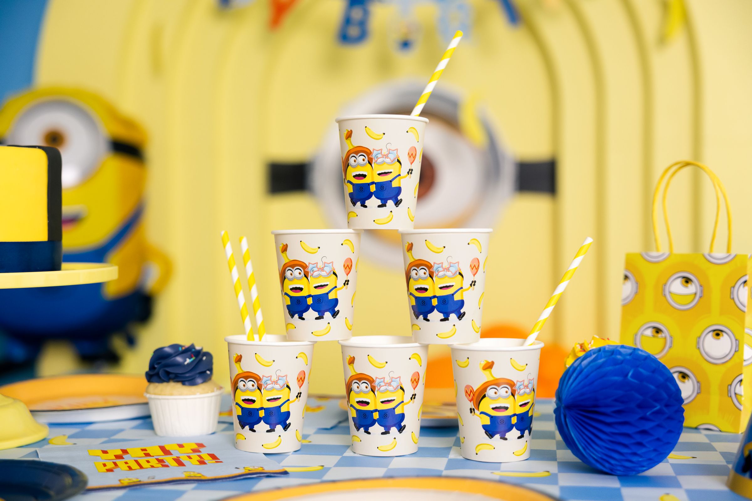 A pyramid stack of six beige Minions paper cups with yellow striped straws, sitting on a blue and white checkered party tablecloth.