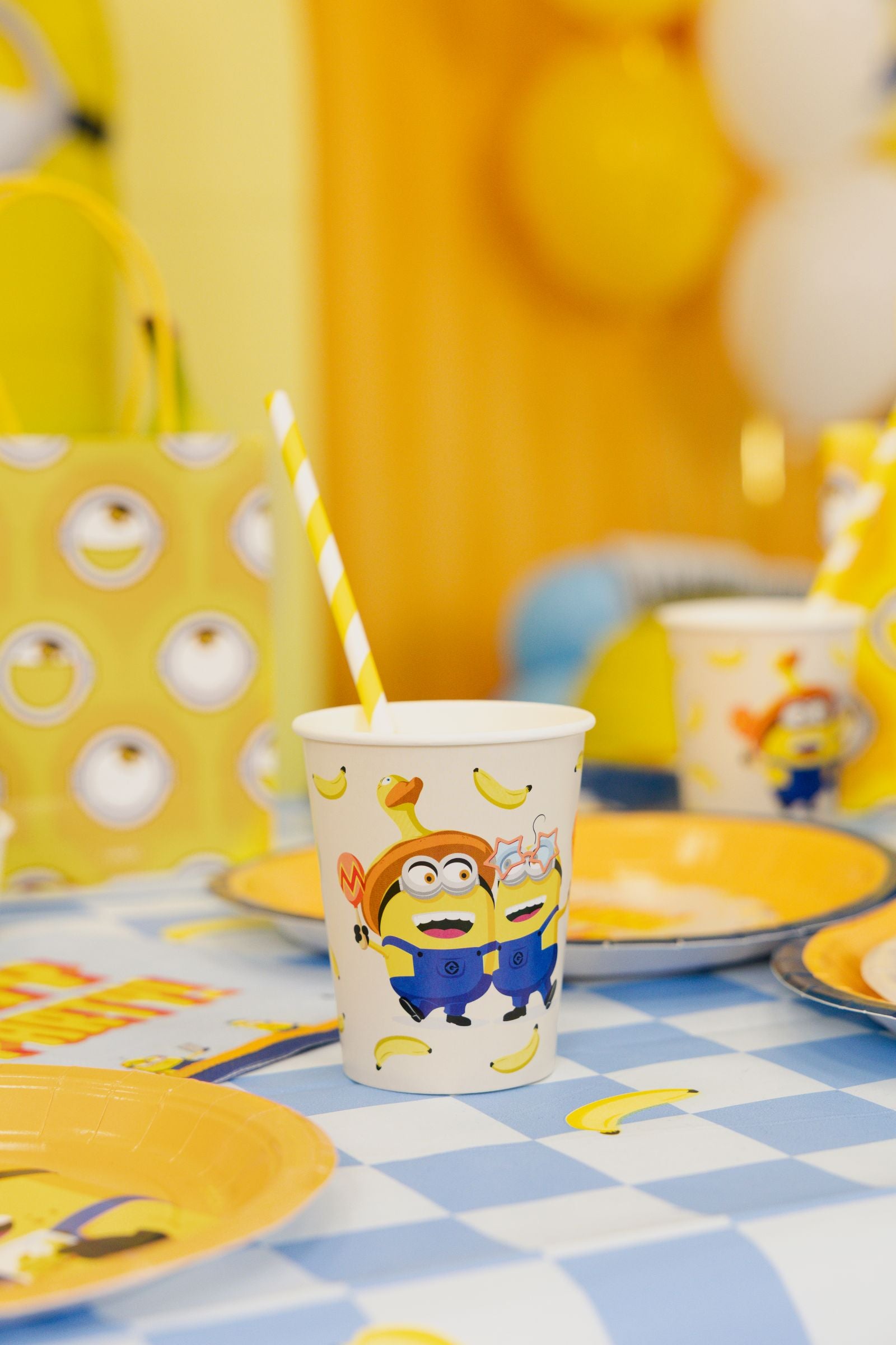 A close-up of a Minions paper cup with a yellow and white striped straw, sitting on a party table next to matching yellow Minions paper plates.