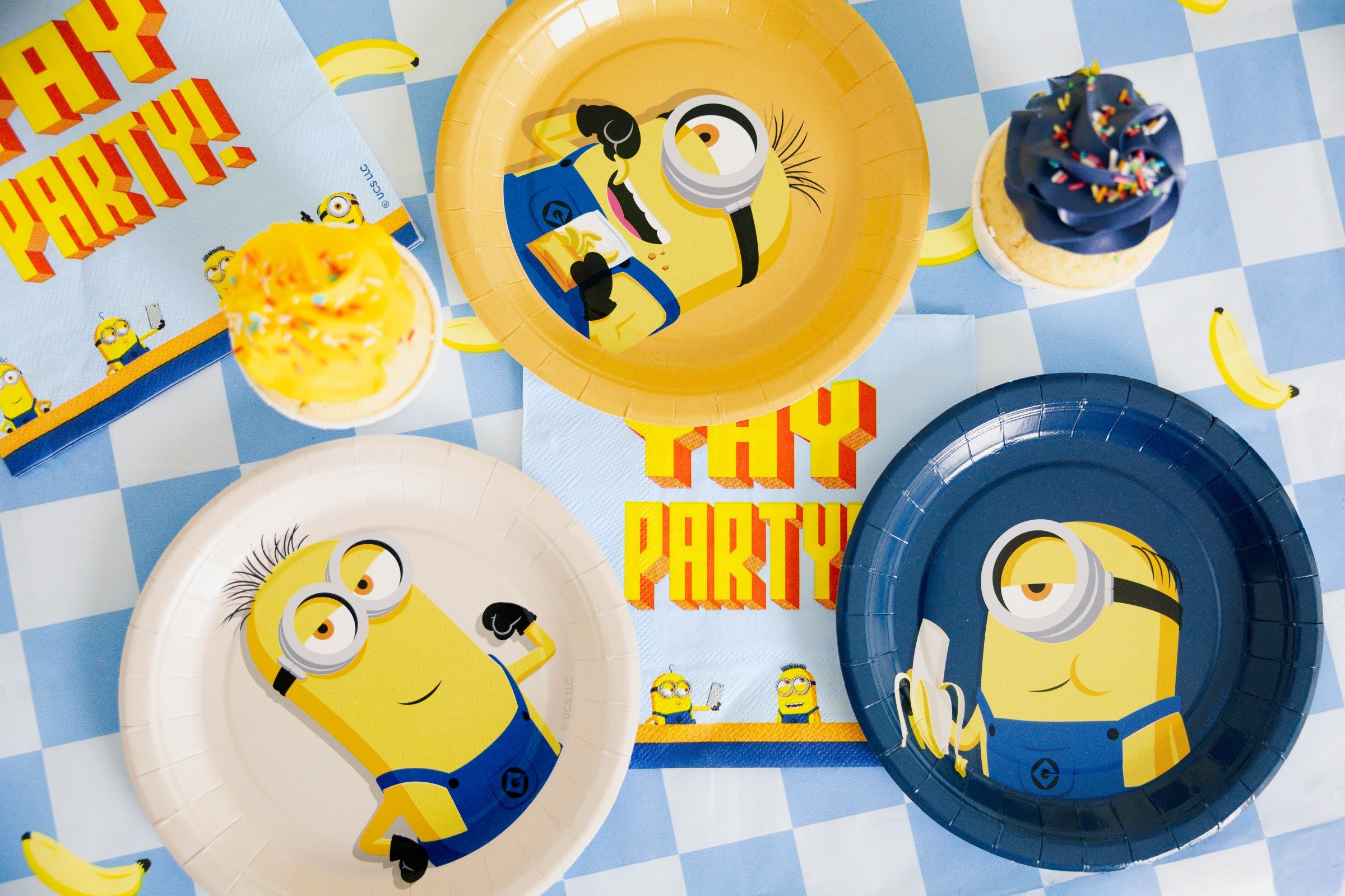 A top-down view of three round Minions paper plates in yellow, beige, and blue, surrounded by matching YAY PARTY napkins and cupcakes on a blue and white checkered tablecloth.