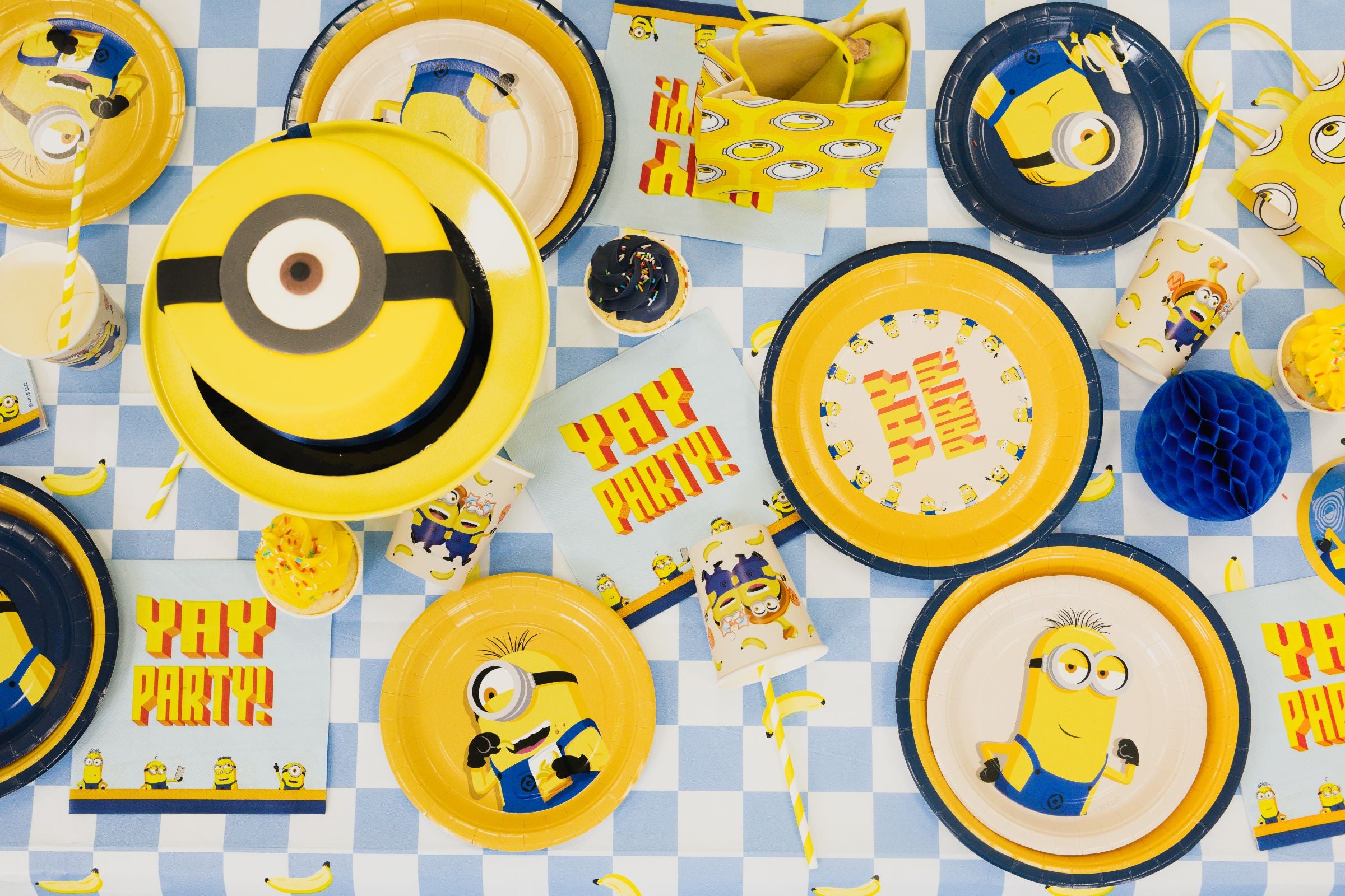A top-down view of a vibrant Despicable Me party table perfectly styled with a blue checkered Minions tablecloth beneath matching plates, cups, napkins, and a Minion cake.
