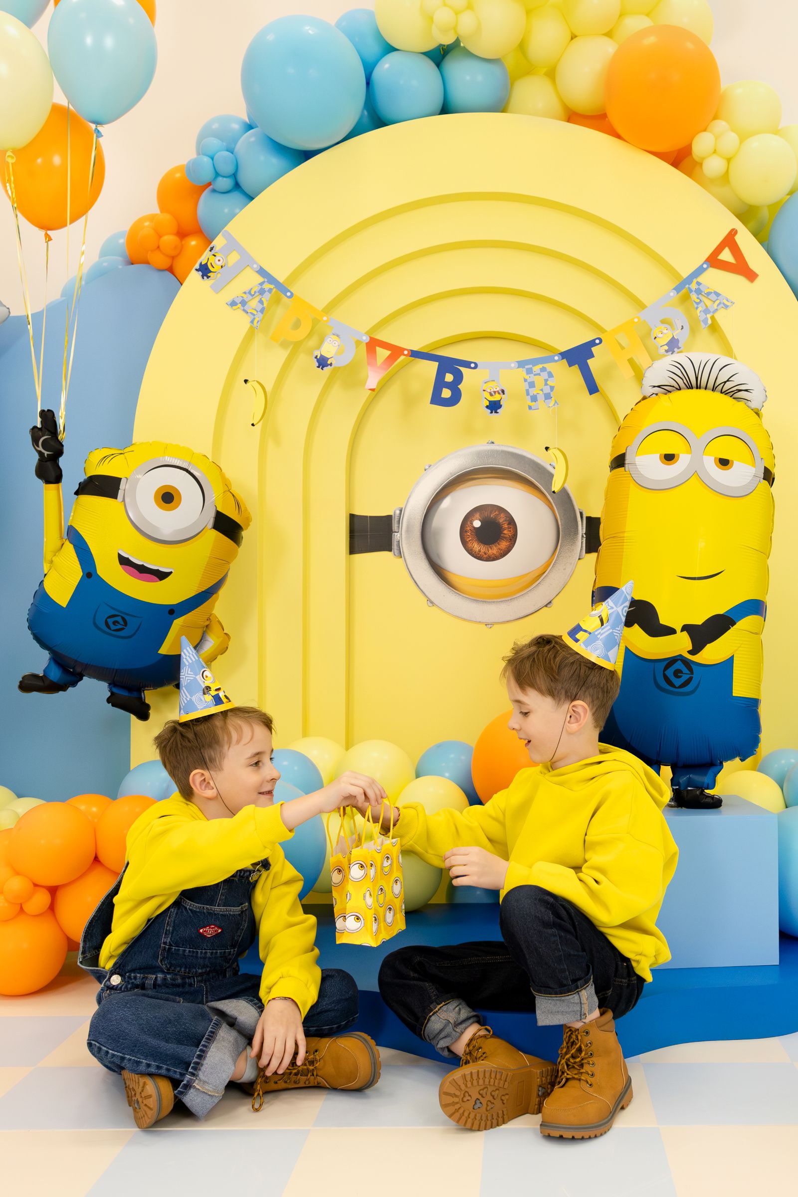 Two happy children sitting on the floor at a Minions-themed birthday party, smiling and holding a yellow Minions paper gift bag together, surrounded by giant Minion foil balloons and a Happy Birthday banner.
