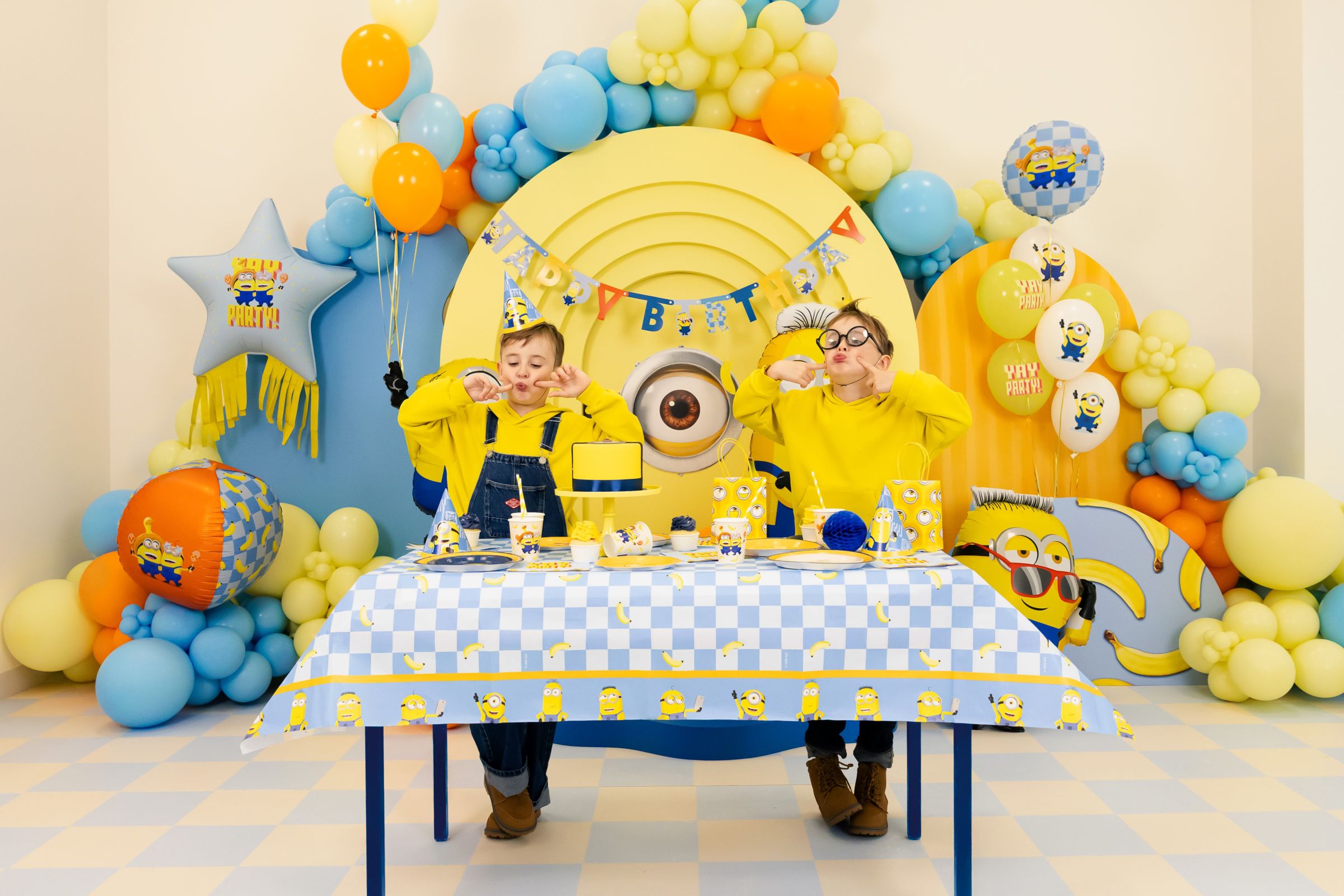Two children sitting at a party table decorated with a blue and white checkered Minions tablecloth, making silly faces in front of a massive Minions-themed backdrop filled with colorful balloons.