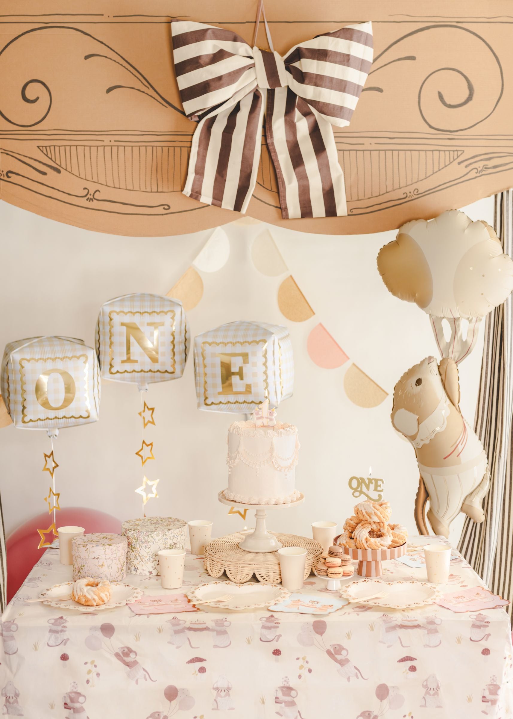 A beautifully styled first birthday dessert table under a cardboard theater arch, decorated with a giant floating mouse foil balloon, a plush bunny, and gingham 'ONE' cube balloons.