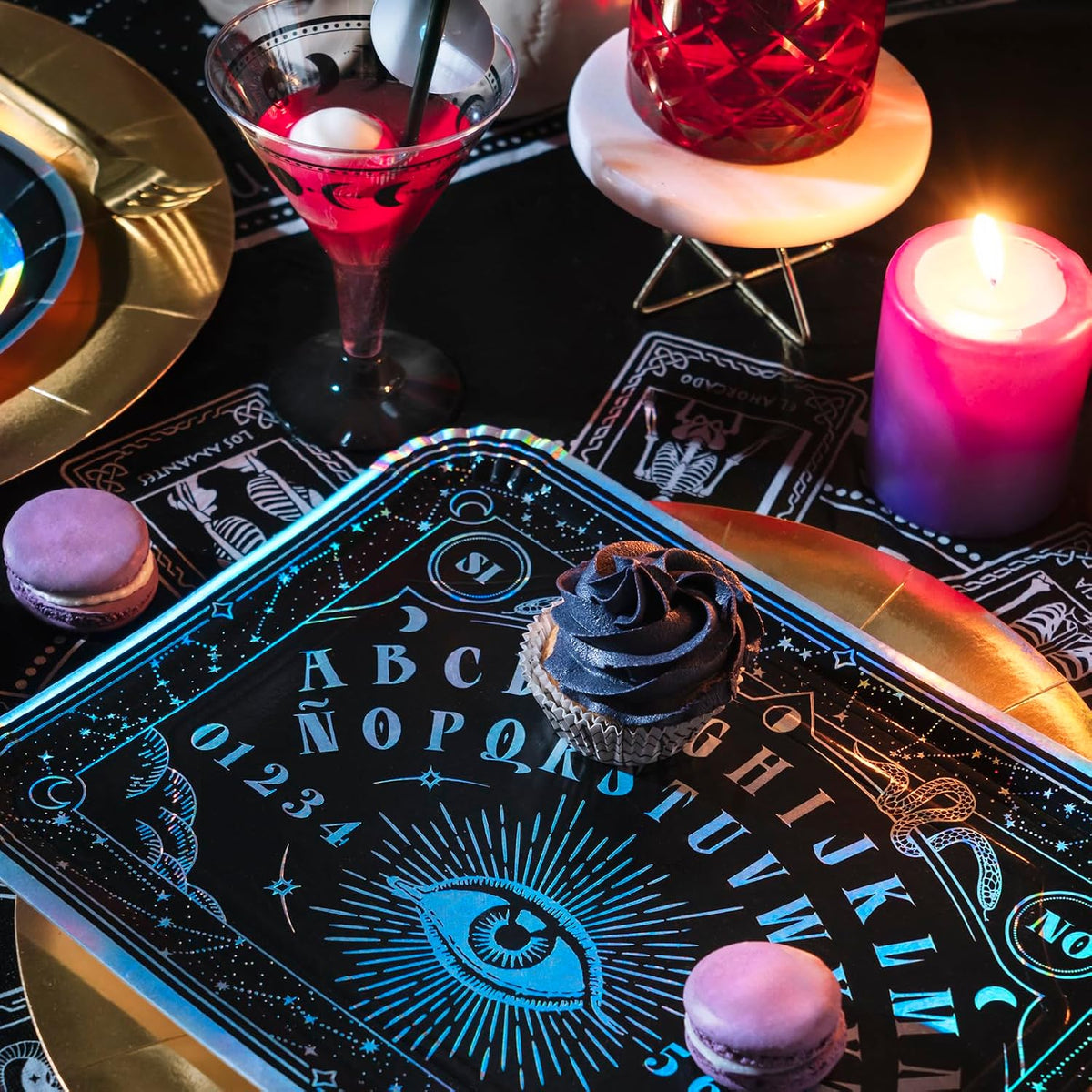 A styled tabletop scene with a black Ouija board tray in the foreground, printed with glowing blue lettering and symbols. A cupcake sits on the board, with macarons, a lit pink candle, a red drink in a martini glass, and gold plates around it on a dark surface.