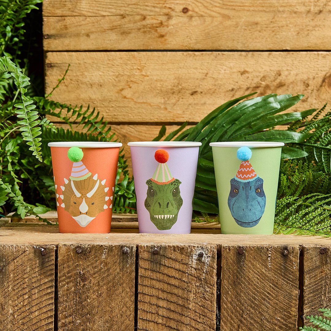 Triceratops, T-Rex, and Raptor paper cups lined up against a wooden backdrop with fern leaves, creating a legendary Jurassic look.