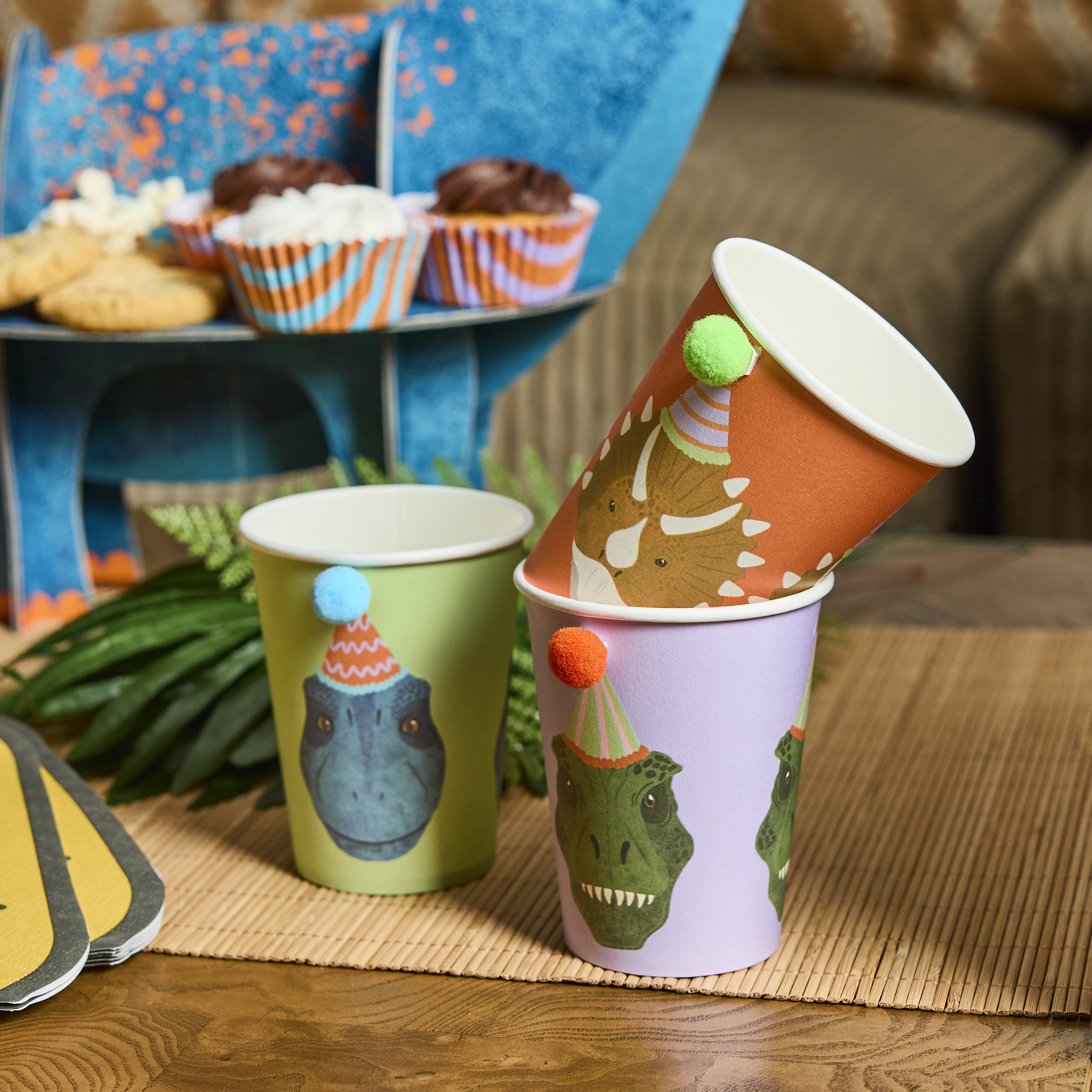Stacked dinosaur party cups on a table next to cupcakes and a blue dinosaur treat stand, ideal for a themed birthday.