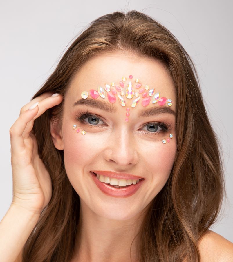 A person smiling directly at the camera, wearing an intricate pattern of pink and iridescent self-adhesive face jewels on their forehead and around their eyes, on a light background.