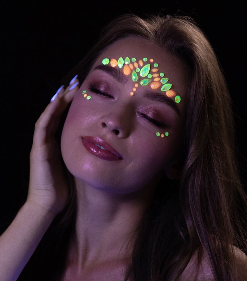 A close-up of a person with eyes closed, wearing glow-in-the-dark face jewels on their forehead and below their eyes, which are emitting a vibrant green and orange glow in a dark setting.