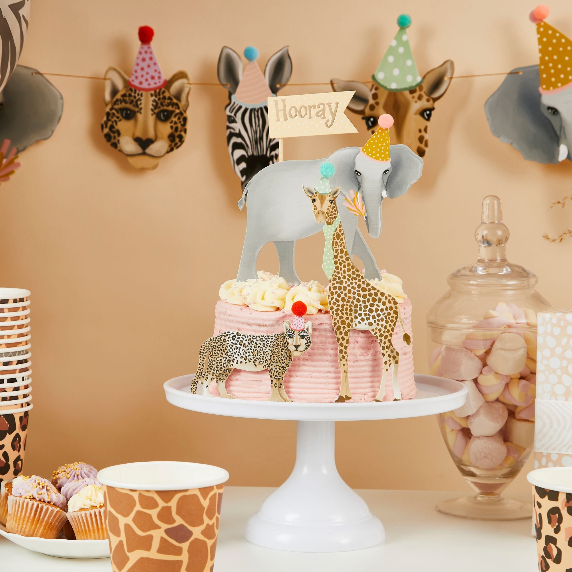 A pink frosted birthday cake topped with safari animal toppers including an elephant, giraffe, and leopard wearing 3D pom-pom hats.