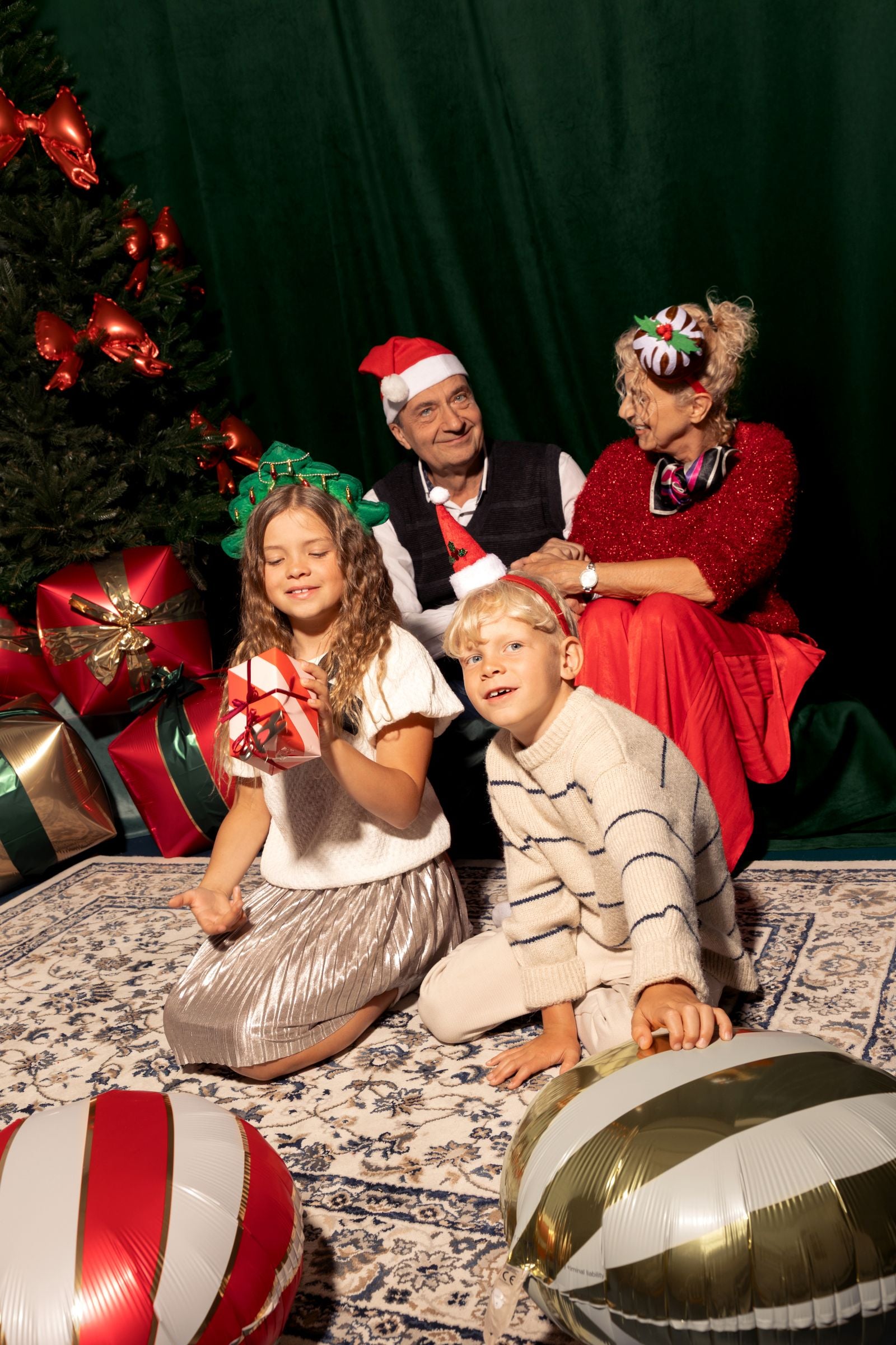 A family poses for a Christmas photo by a tree; the man in the back is wearing a mini Santa hat headband.