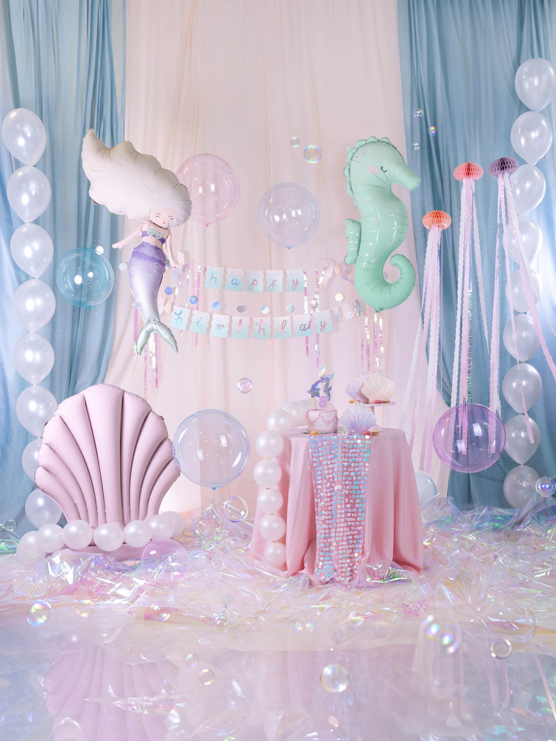 A whimsical mermaid-themed party setup with a large pink seashell foil balloon, a mermaid balloon, a seahorse balloon, and a "Happy Birthday" banner against blue and white draped fabric, with clear balloons and iridescent floor decor.