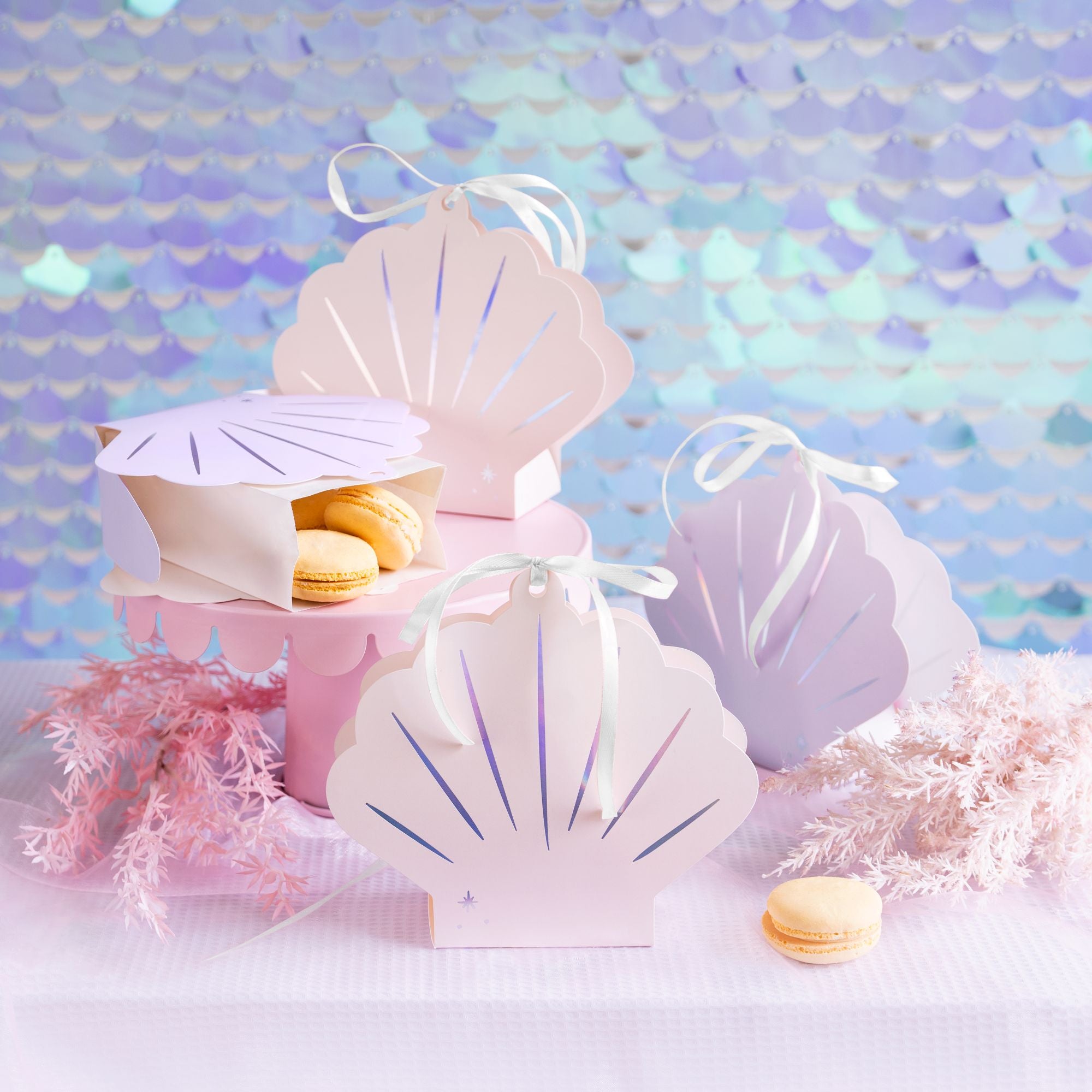 A close-up of several iridescent seashell-shaped snack boxes arranged on a table with a shimmering purple and pink mermaid scale backdrop. One box is open and contains yellow macarons, while others are closed and decorated with ribbons.