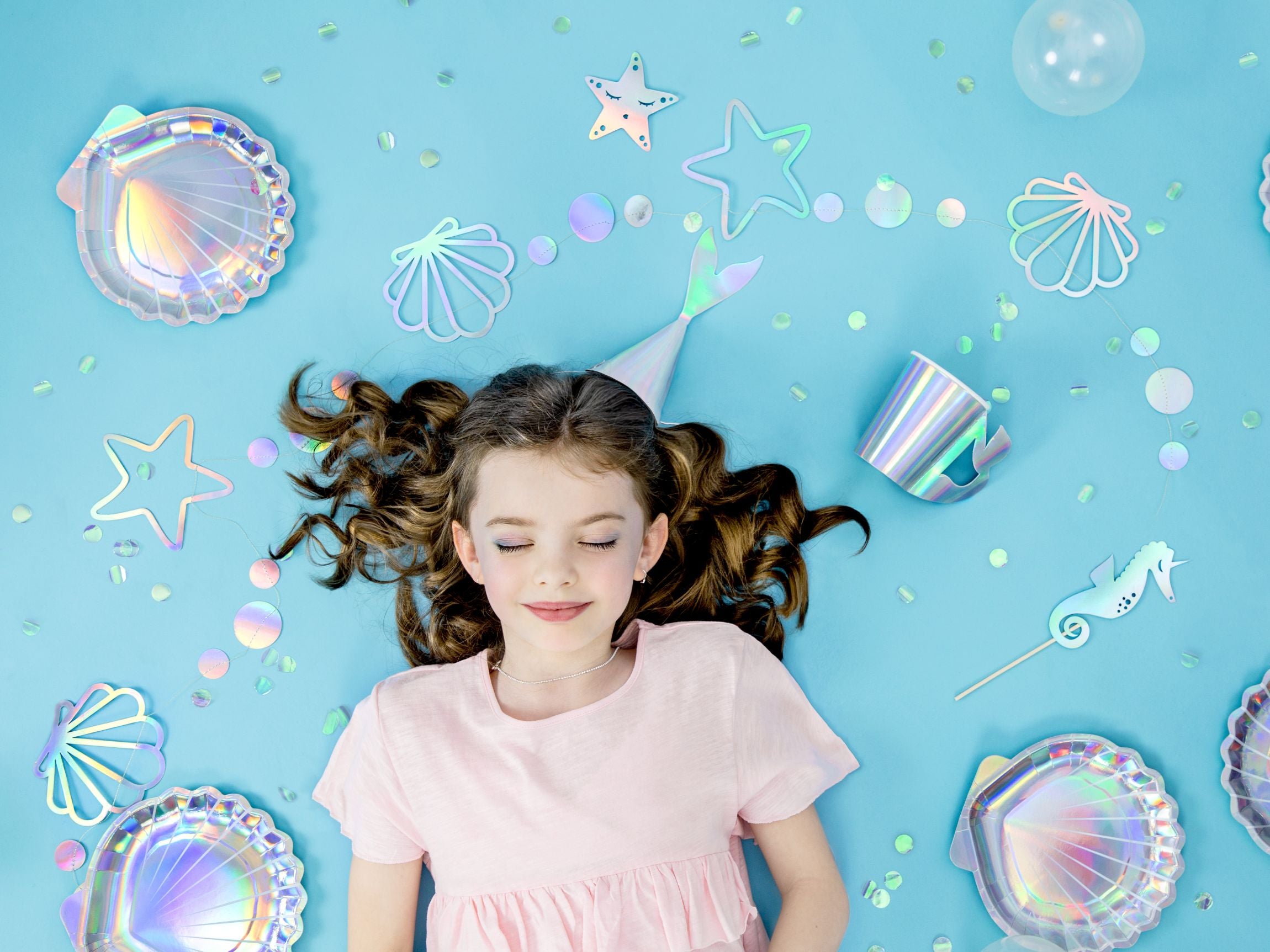 An overhead view of a festive mermaid-themed party arrangement on a light blue background, showing a child with closed eyes lying amidst various iridescent seashell plates, starfish cutouts, a mermaid tail decoration, a matching cup, and scattered confetti and clear balloons.