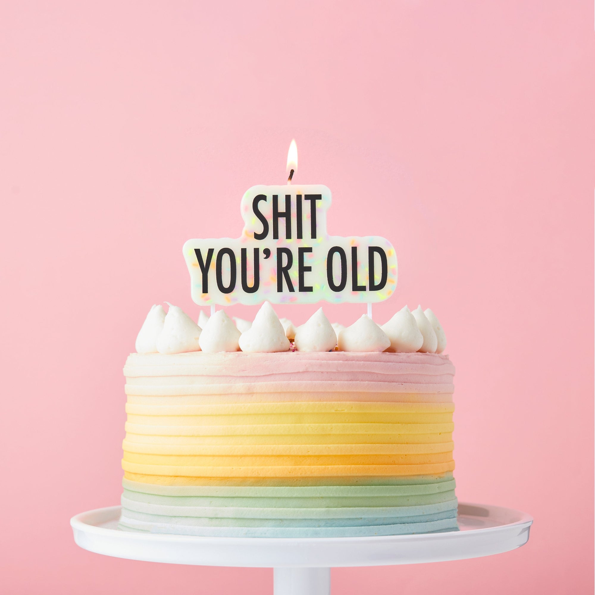 A white novelty birthday cake candle shaped like a text bubble with colorful sprinkles, featuring the words "SHIT YOU'RE OLD" in bold black letters and two plastic picks at the bottom.