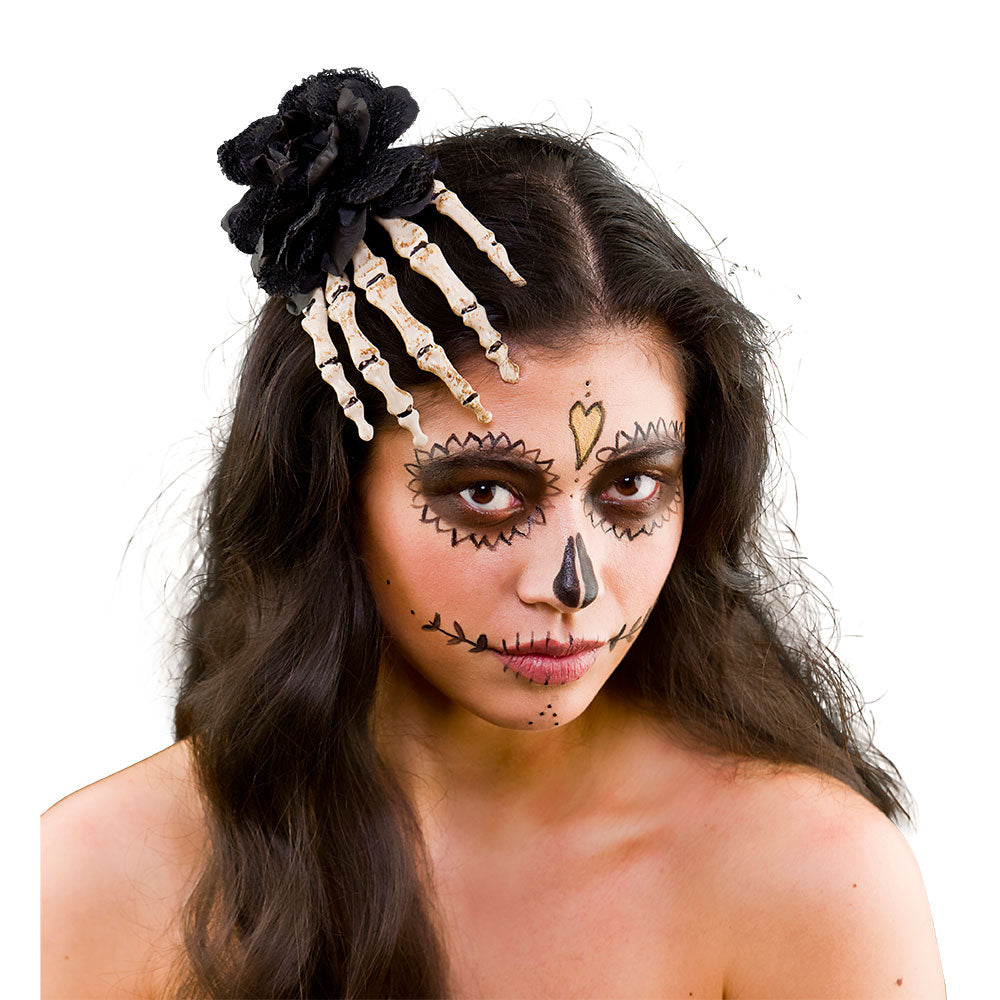 A close-up of a person with long dark hair and Dia de los Muertos (sugar skull) style makeup, wearing a black lace rose and bone-colored skeleton hand hair clip as a headpiece.