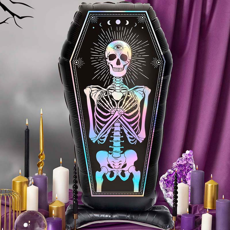 An inflatable black coffin-shaped balloon or decoration featuring a colorful pastel rainbow skeleton with an eye on the skull, displayed on a table with purple and gold candles, crystals, and a purple draped backdrop.