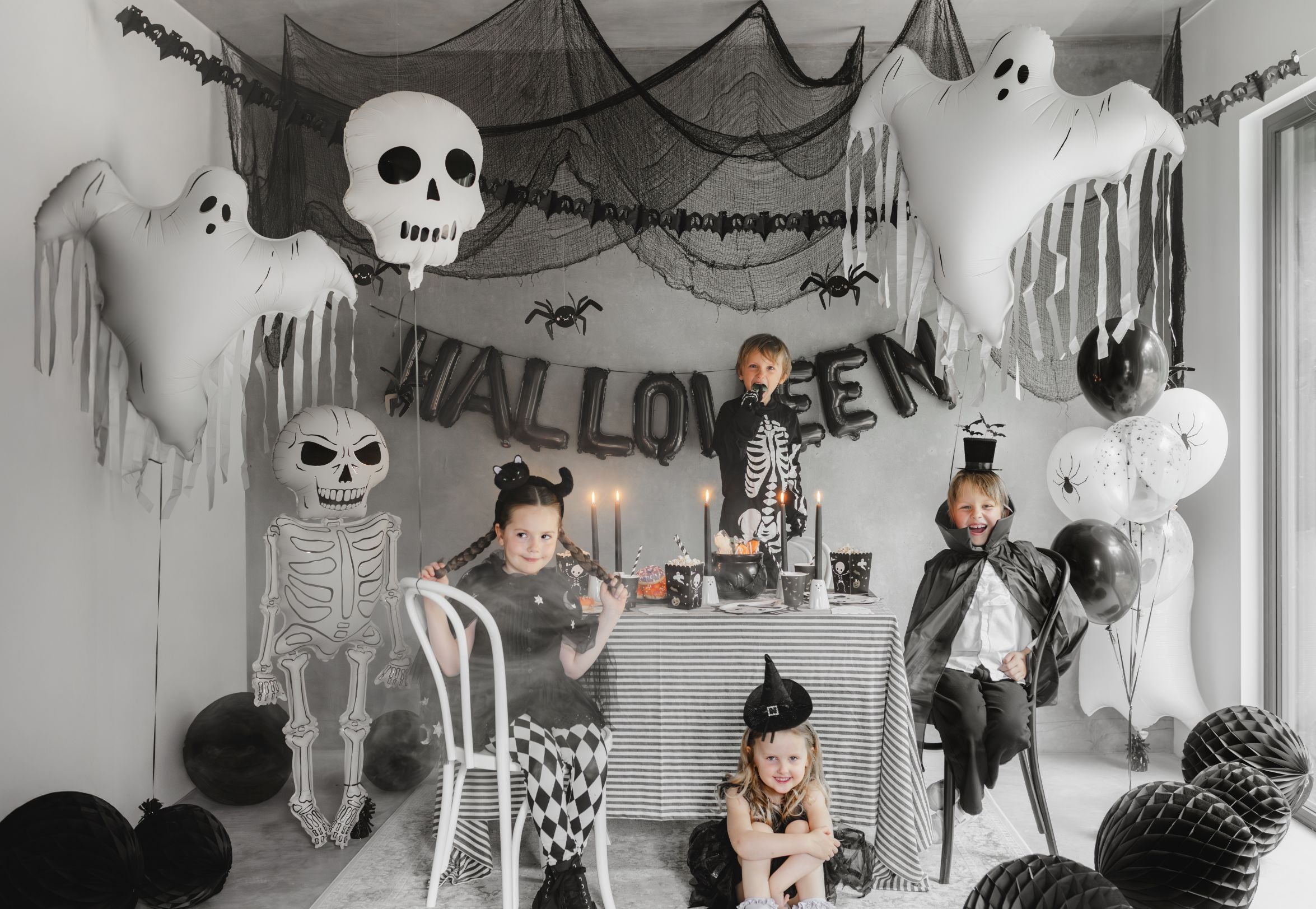 A lively Halloween party scene featuring children in various costumes (skeleton, vampire, witch) around a decorated table. The room is adorned with black netting, spider decorations, ghost balloons, and prominently, a large white skull foil balloon hanging in the center.