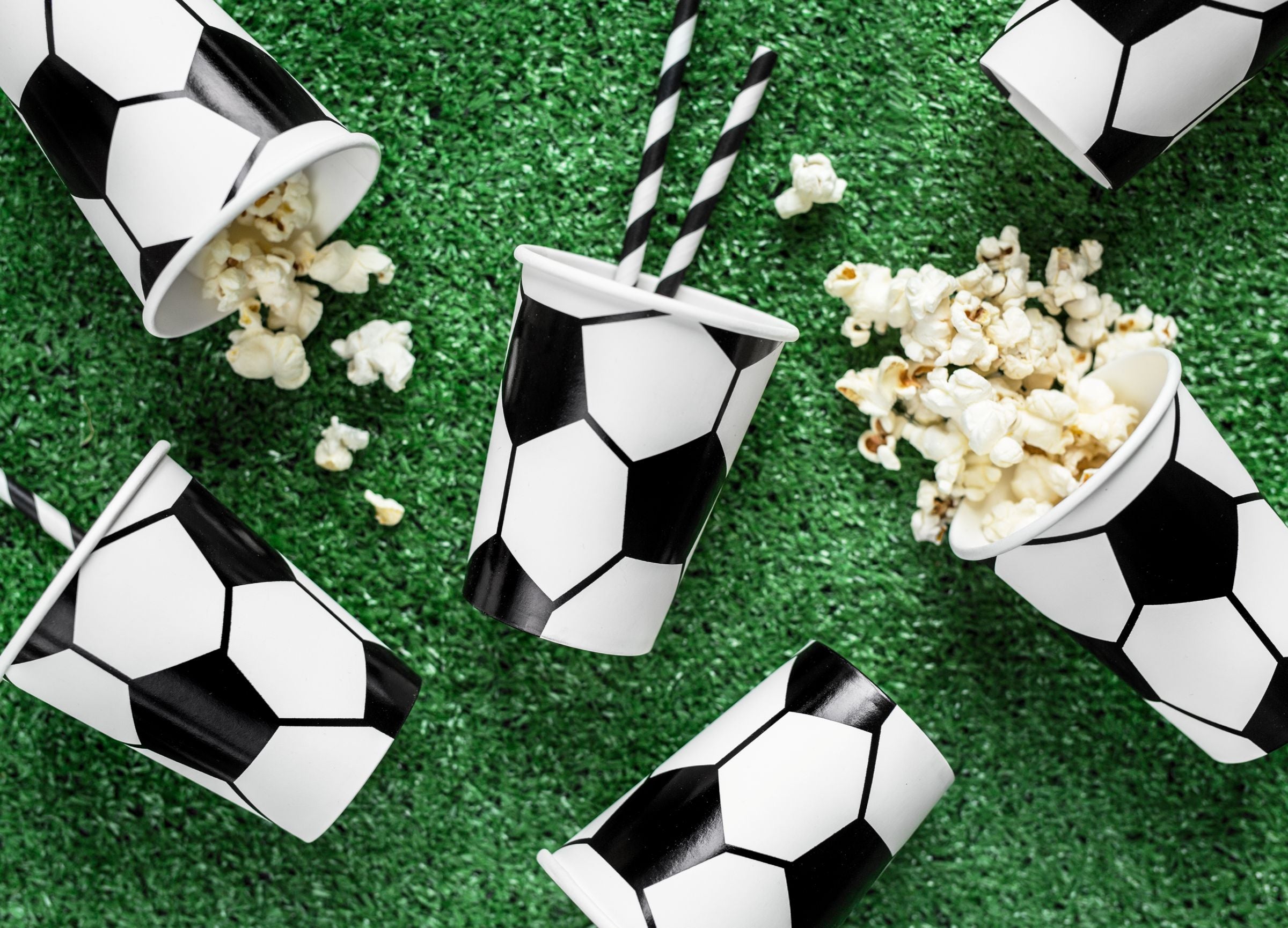An overhead view of multiple black and white soccer ball patterned paper cups scattered across a green turf-like surface, with some cups filled with popcorn and others containing striped straws.