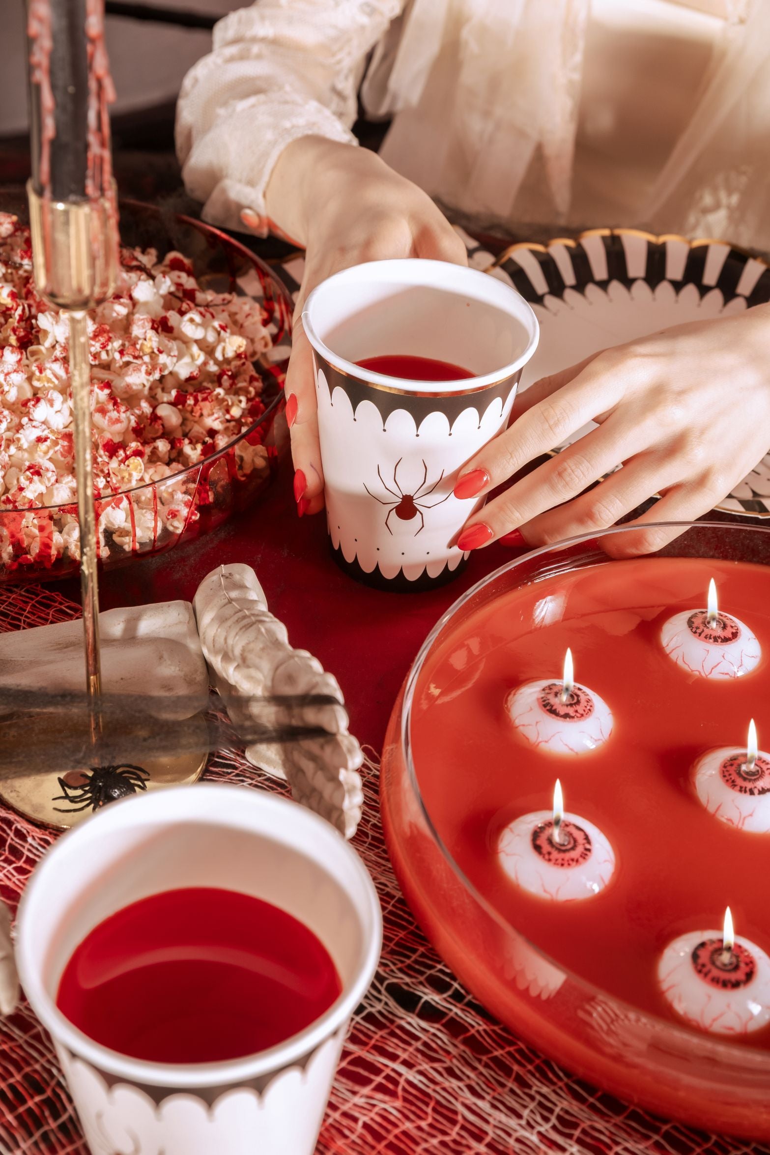 A close-up of a Halloween party table setup with a red tablecloth. Hands with red nail polish hold a white spider-themed cup filled with red liquid. Other items include a bowl of popcorn, a decorative skeleton hand, a punch bowl with "eyeball" candles floating, and a black and white spider-themed plate.