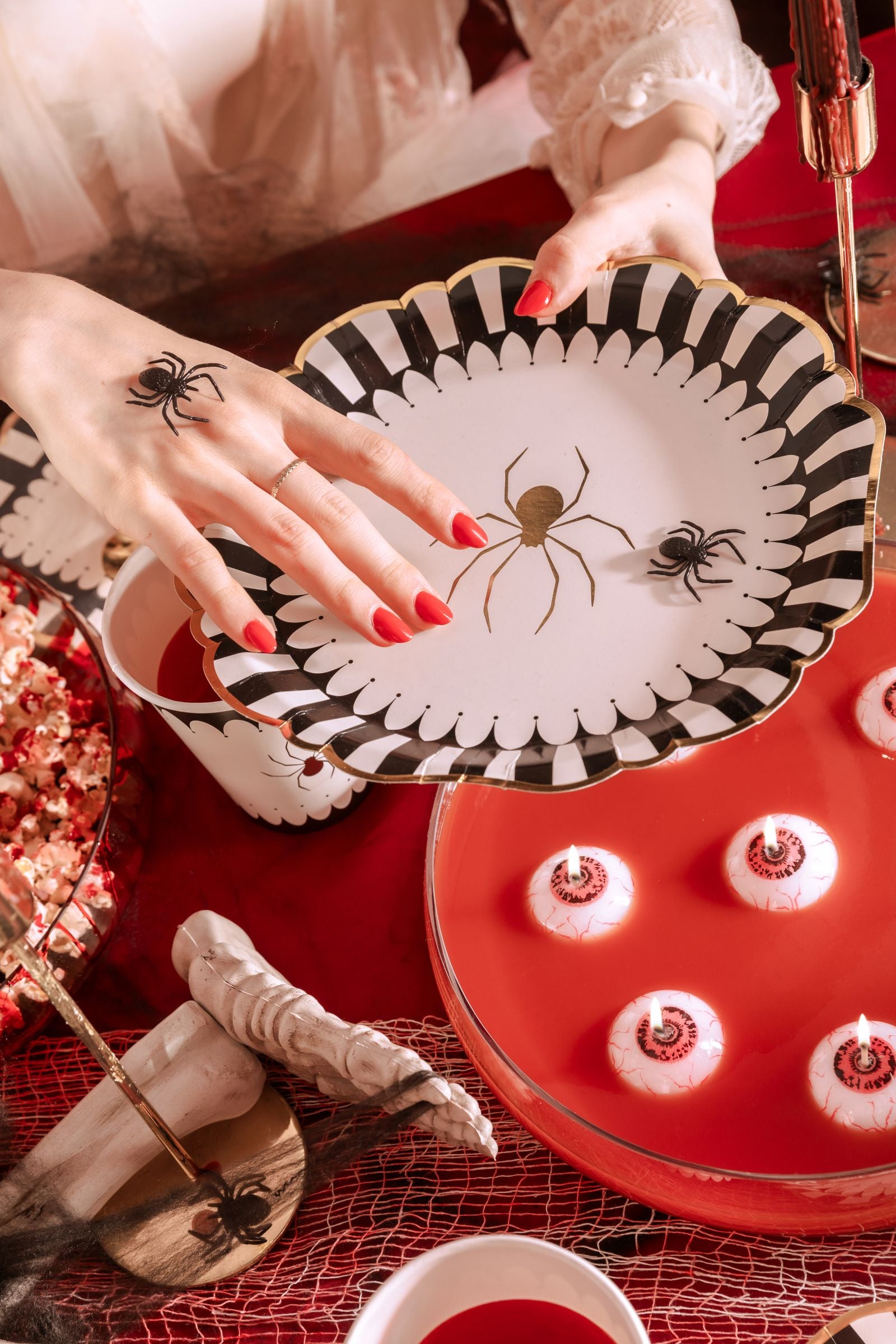 An overhead view of a Halloween-themed table decorated with a red tablecloth and black netting. Hands with red nail polish are placing or holding a white plate with a black and white scalloped edge and a gold spider design, above a red punch bowl with floating "eyeball" candles. Other decorations include a skeleton skull and bones, popcorn, and themed cups.