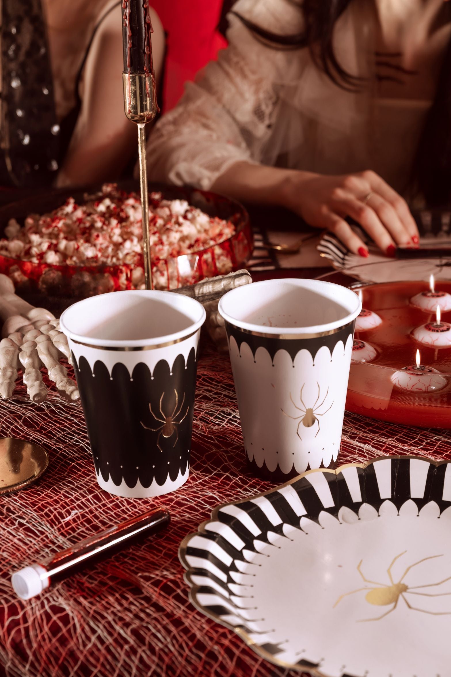 A close-up of a Halloween party table setup with a red tablecloth. Two spider-themed cups (one black, one beige, both with gold spiders) are prominent, along with a striped plate, a skeleton hand, and a bowl of popcorn with fake blood.