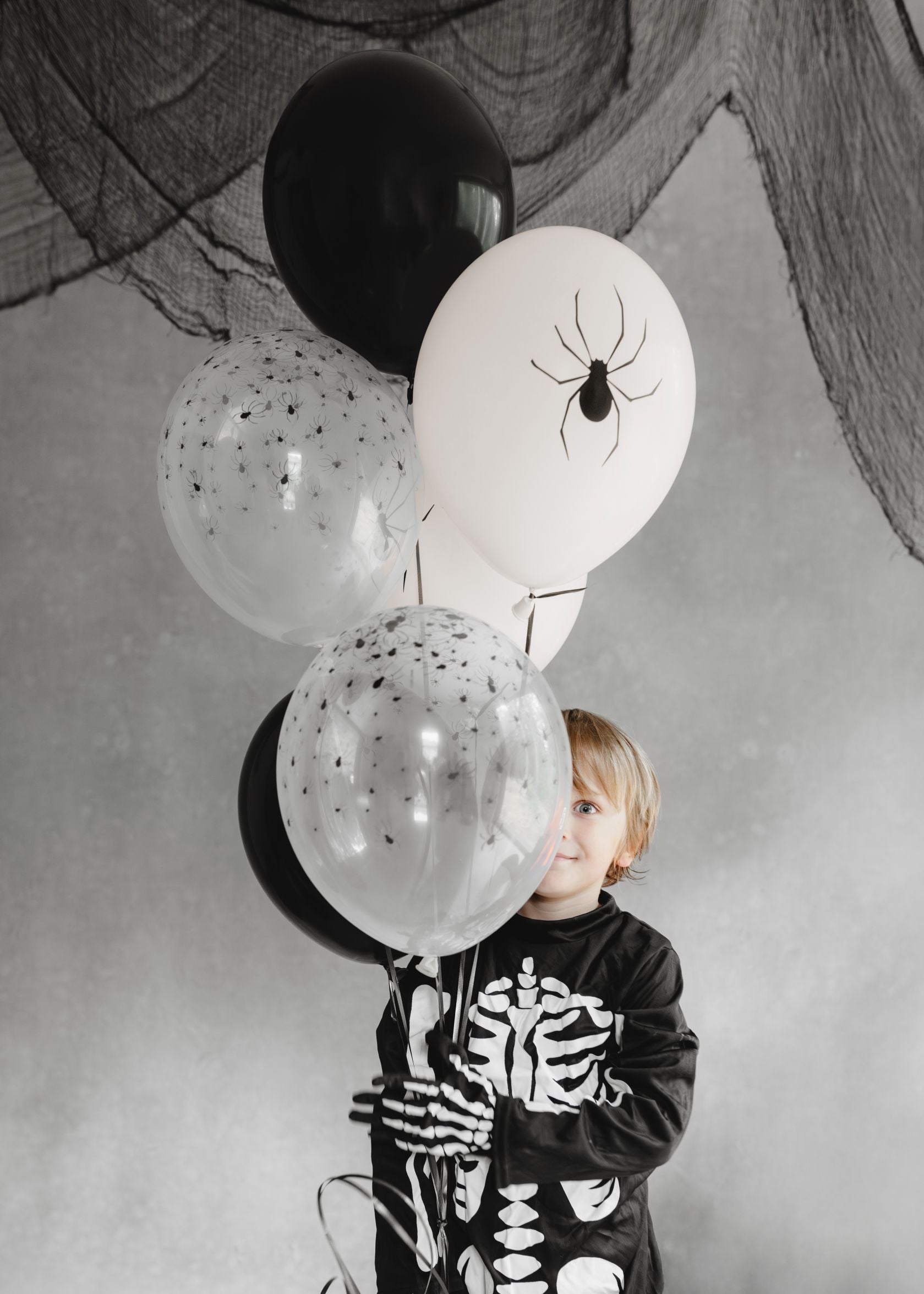 A child dressed in a skeleton costume, holding a bunch of Halloween-themed balloons, including black, white with a large spider, and clear with tiny spiders, against a grey wall draped with black netting.