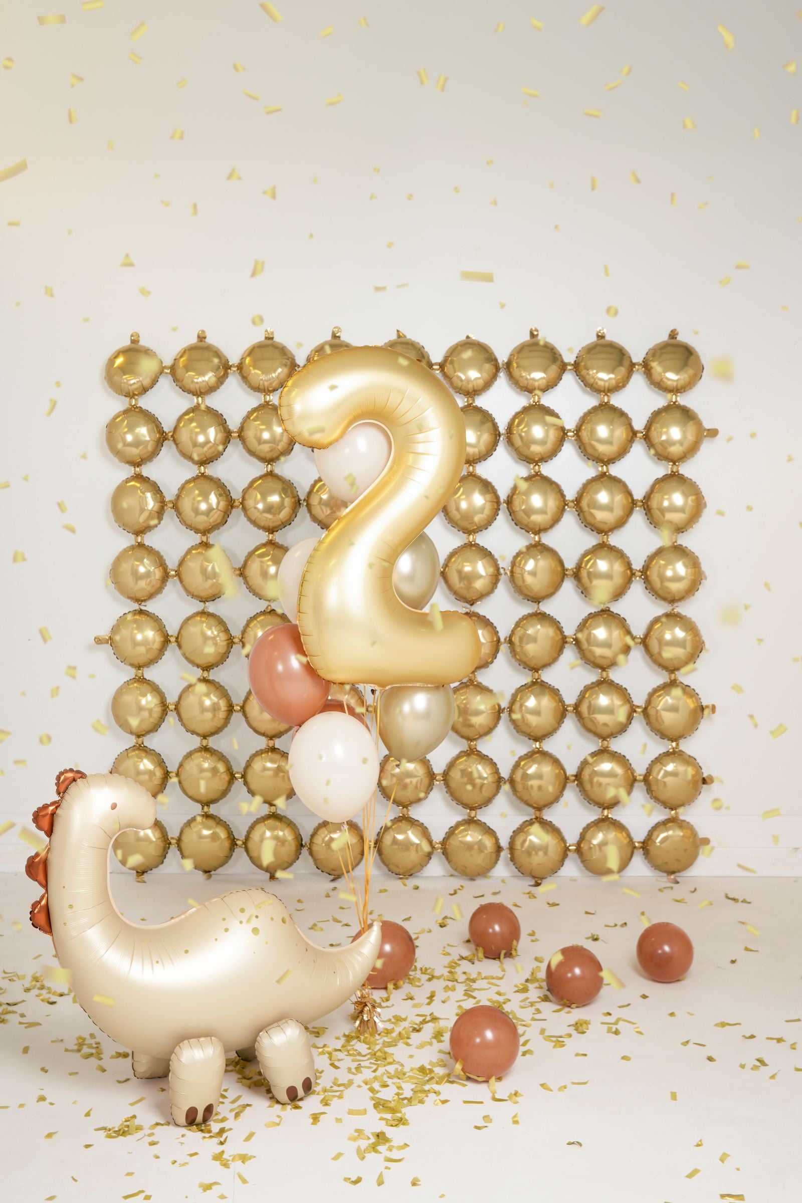 A party scene with a large cream-colored brontosaurus foil balloon on the floor, surrounded by small brown balloons and gold confetti. In the background is a wall decorated with a grid of gold foil balloons and a large gold number "2" balloon.